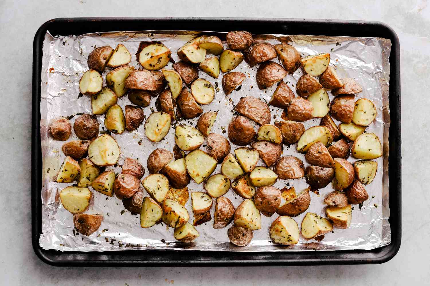 A sheet pan of roasted red potatoes with golden crispy edges, rosemary leaves, and browned garlic on a parchment-lined pan in a bright home kitchen