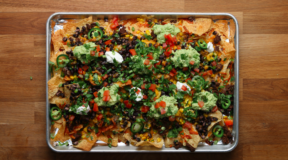 A sheet pan of stacked nachos arranged in neat rows like little books, with melted cheese, pickled jalapeños, diced tomatoes, and cilantro on a parchment-lined pan on a warm kitchen counter
