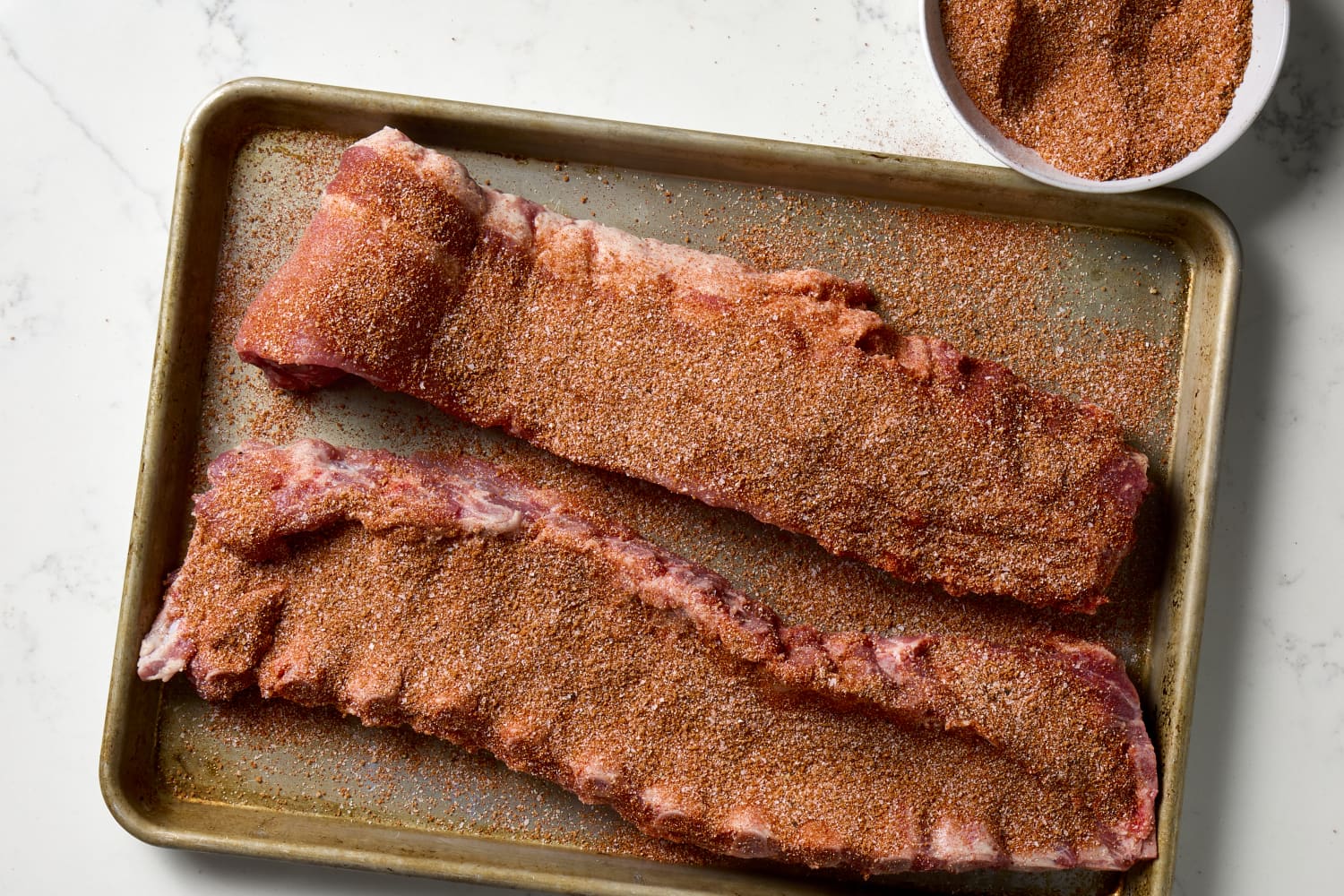 A sheet pan on a kitchen counter with baby back ribs being seasoned heavily with a reddish-brown spice rub, with small bowls of spices nearby