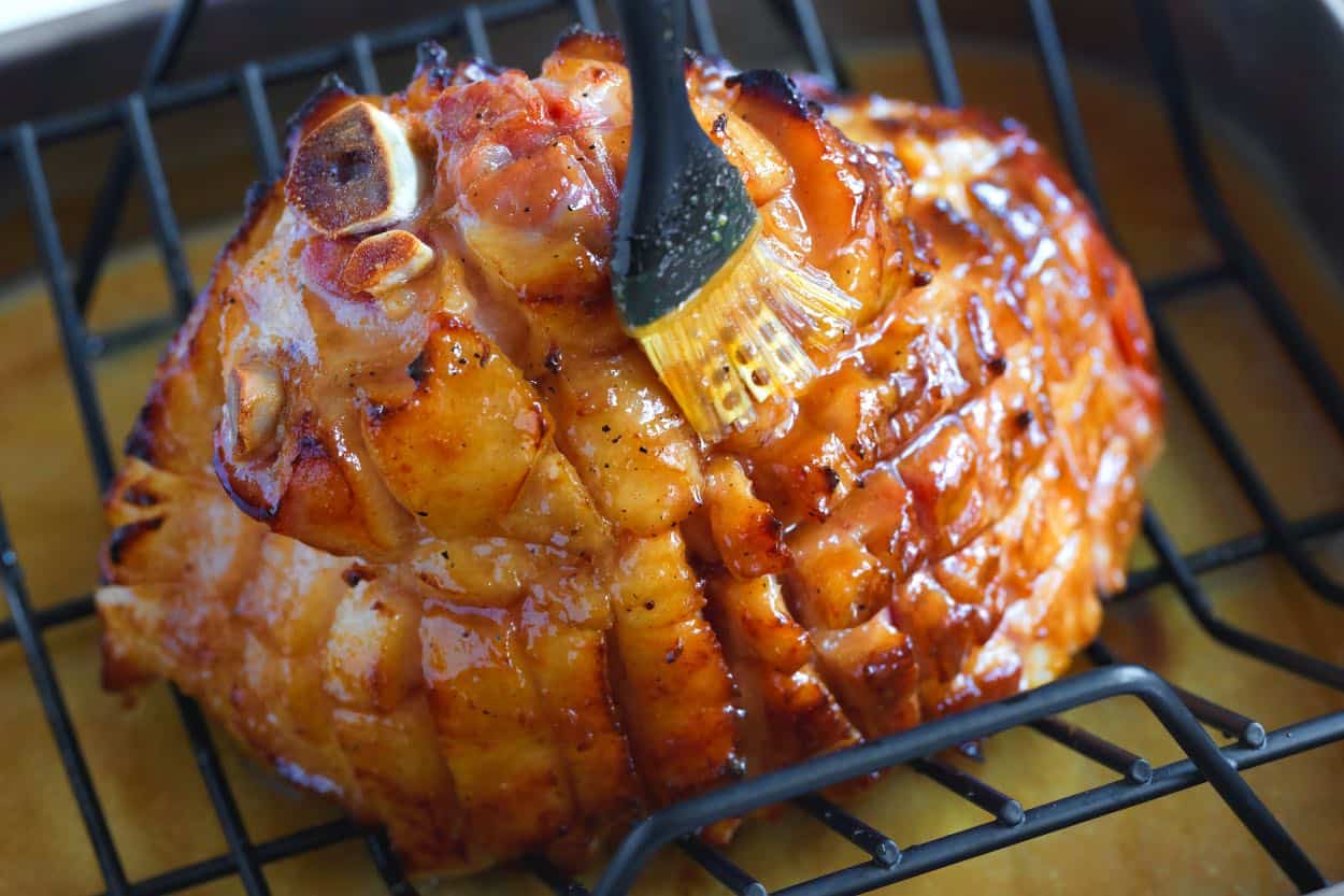 A silicone basting brush applying glossy honey glaze over a scored ham in a roasting pan