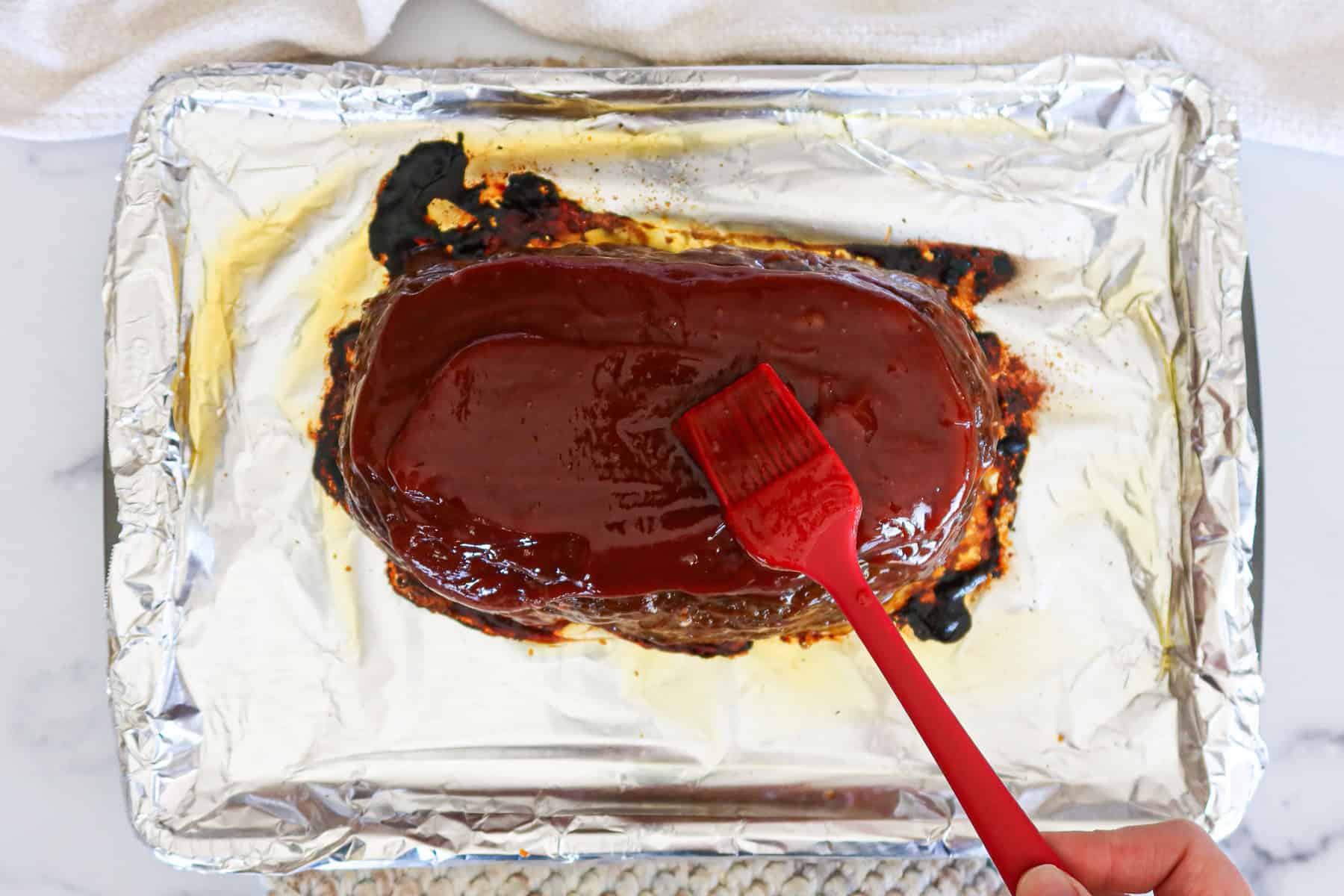 A silicone brush spreading glossy meatloaf sauce over an uncut meatloaf in a baking pan