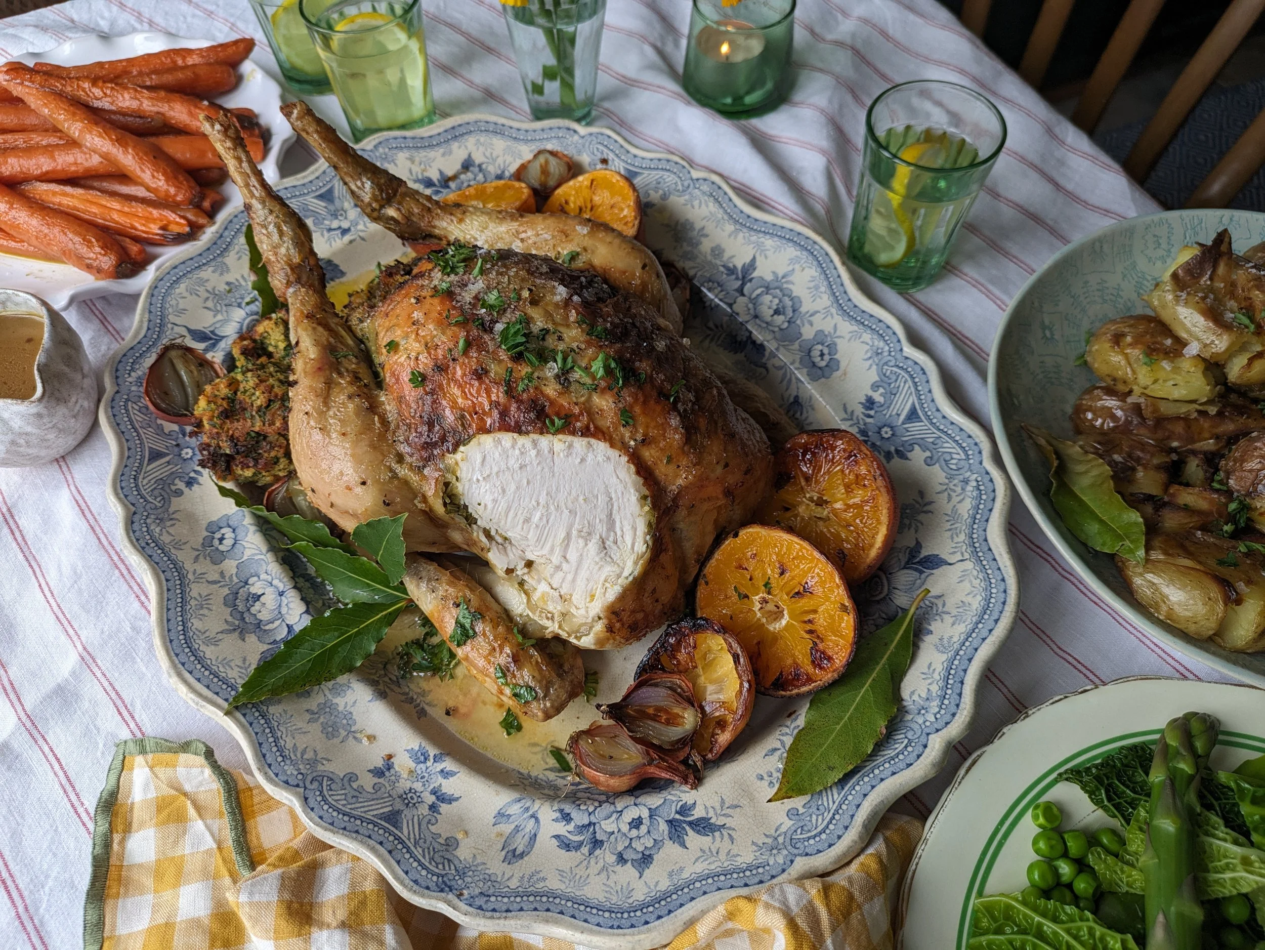 A simple Easter table setting with a platter of roast chicken, scattered herbs, and soft natural light coming through a window