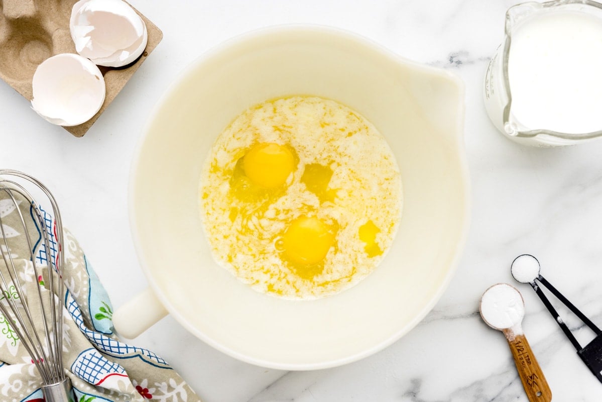 A simple ingredient setup on a kitchen counter with a bowl of cornmeal, a small bowl of flour, buttermilk in a measuring cup, eggs, melted butter, and baking powder