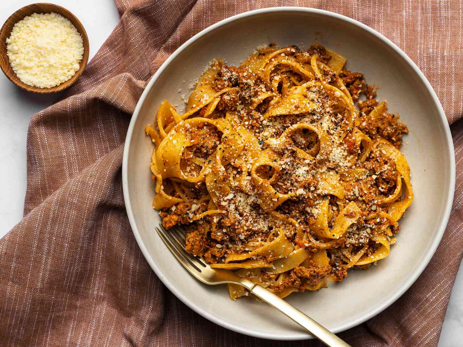 A single bowl of pasta bolognese with glossy red meat sauce, grated Parmesan, and a few basil leaves on a rustic wooden table with warm kitchen lighting