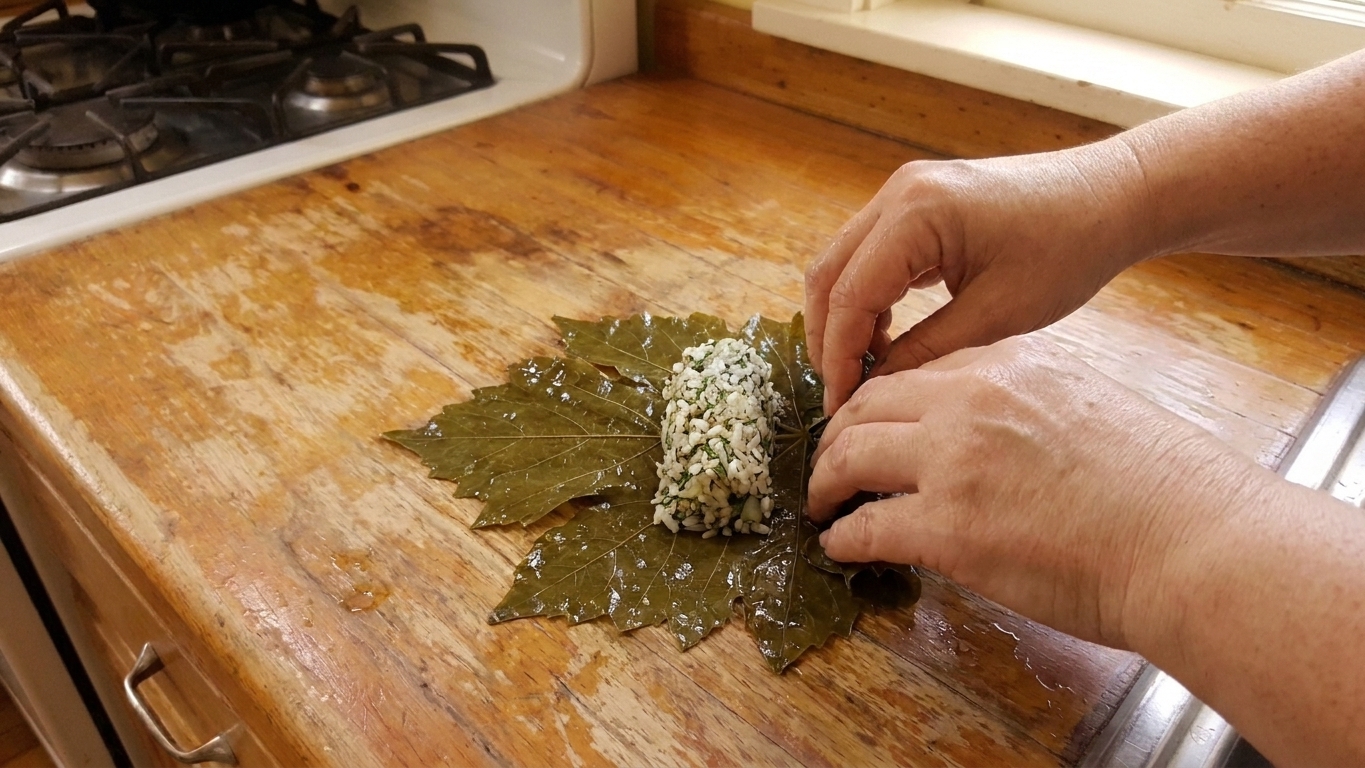 A single grape leaf on a cutting board being rolled around a small mound of herbed rice filling, hands folding the sides inward