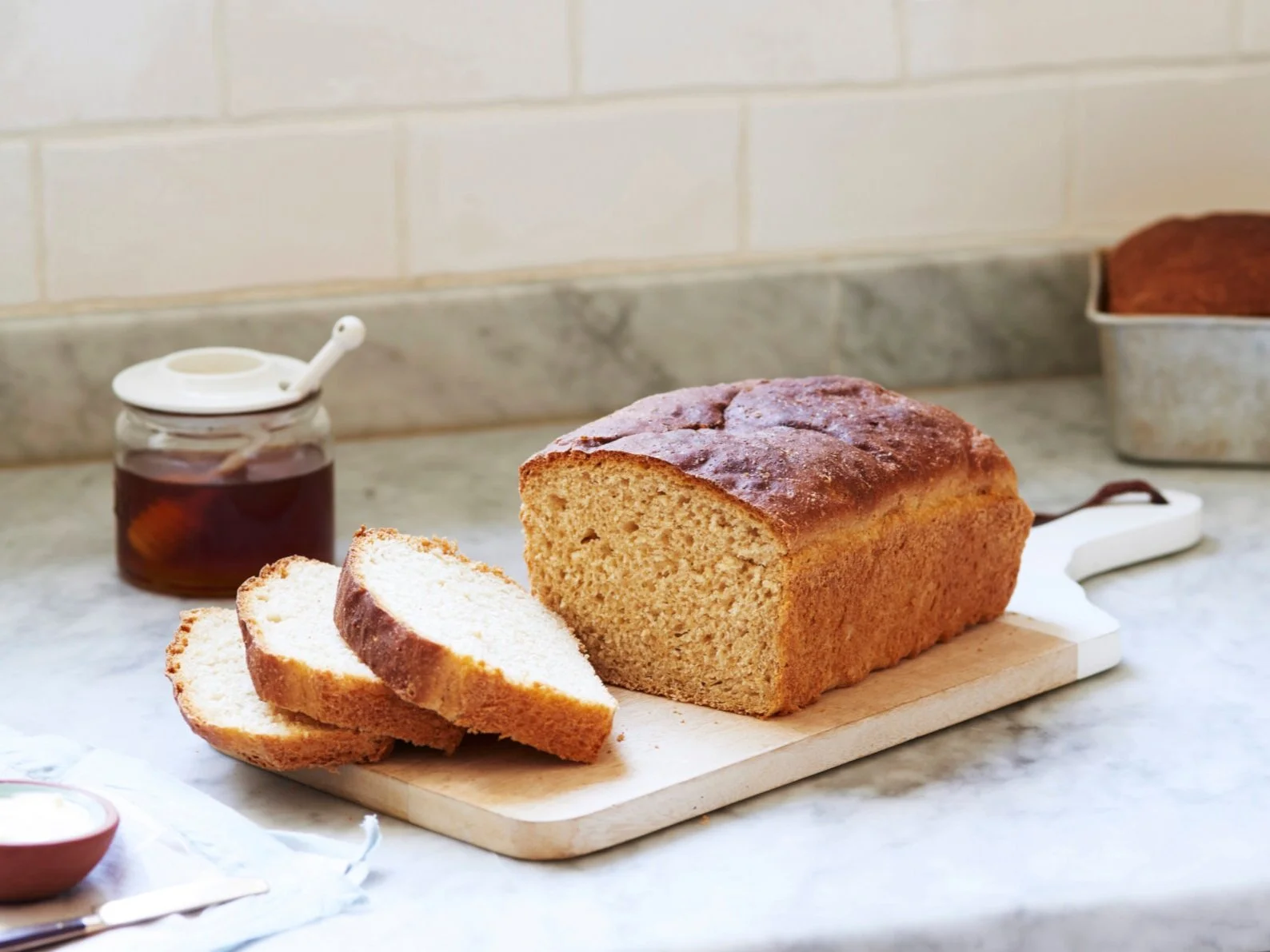 A single loaf of honey wheat bread with a golden-brown crust, sliced to show a soft tan crumb, resting on a wooden cutting board on a bright kitchen counter