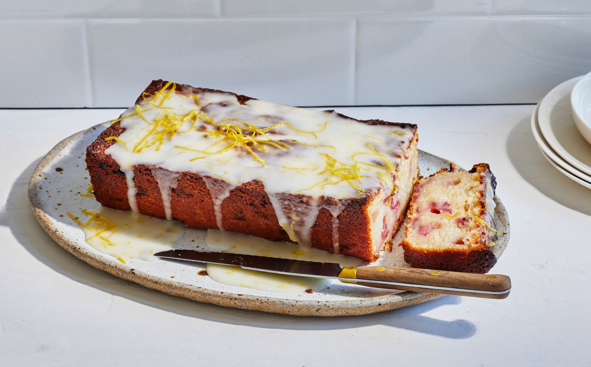 A single loaf of strawberry bread on a wooden cutting board, drizzled with lemon glaze, with a few sliced strawberries scattered nearby on a bright kitchen counter