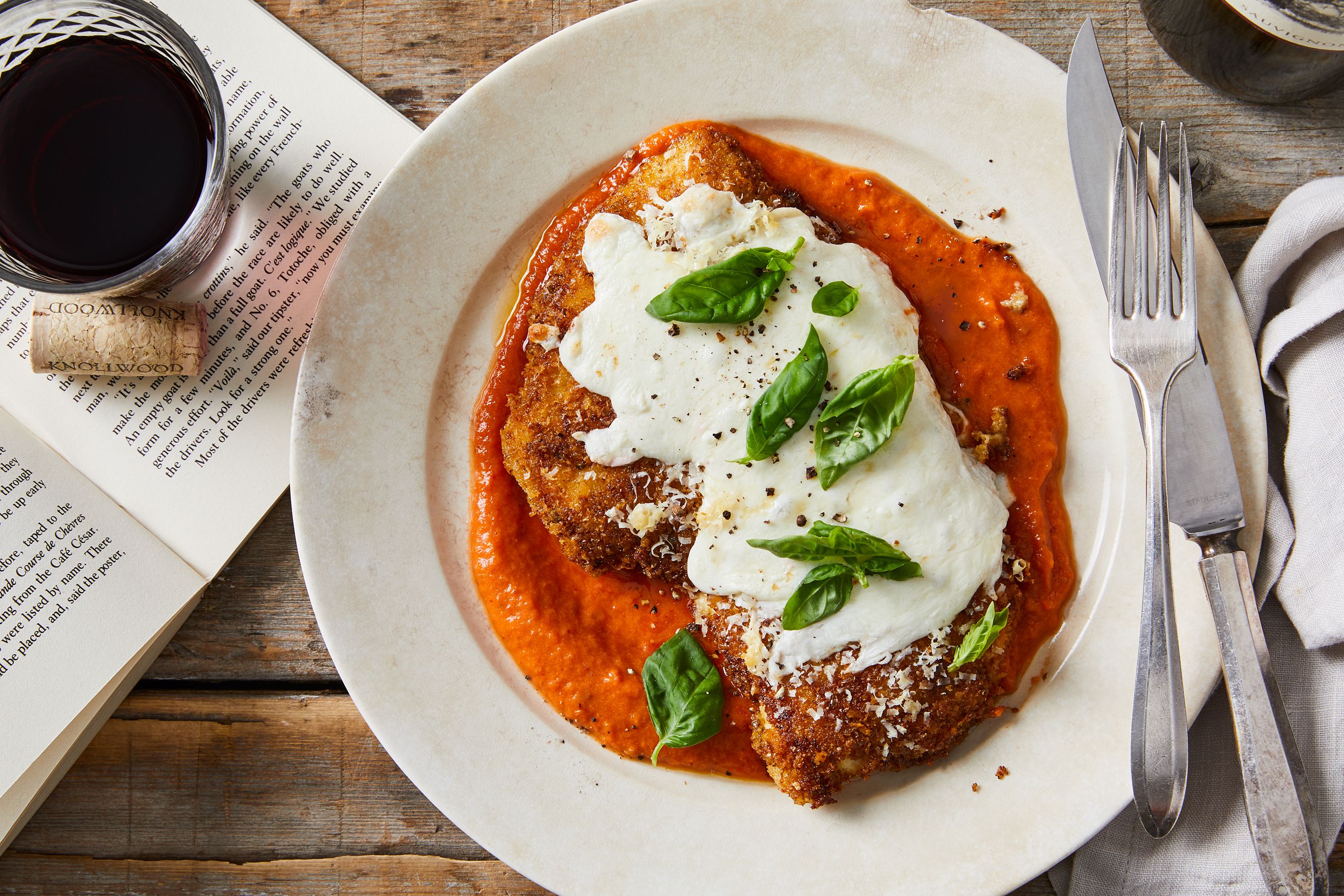 A single plate holding crispy chicken parmigiana topped with melted mozzarella and marinara, with fresh basil and grated Parmesan on a wooden table under warm kitchen lighting