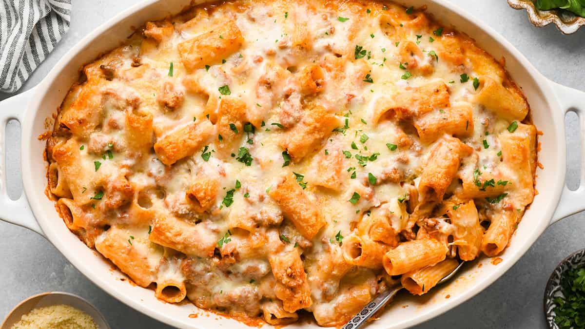 A single portion of baked mostaccioli on a plate with melted cheese and red sauce, a fork cutting into it on a kitchen table