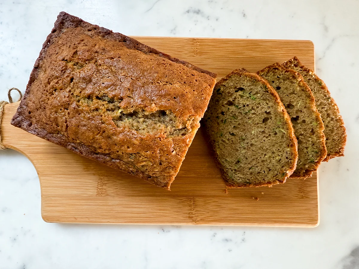 A single real photograph of a sliced loaf of homemade zucchini bread on a wooden cutting board with visible green zucchini flecks, warm natural kitchen light