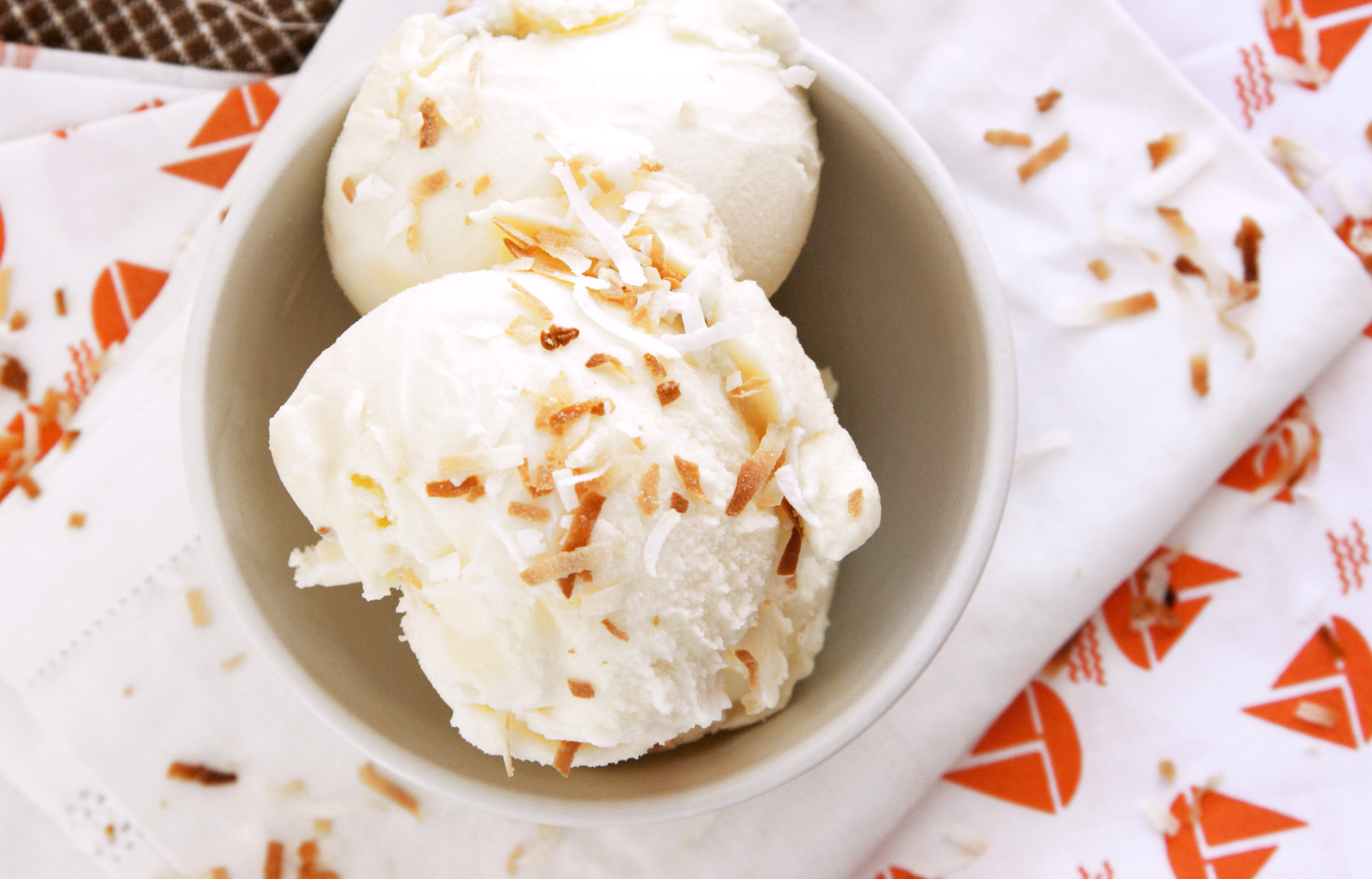 A single scoop of creamy coconut ice cream in a white bowl with toasted coconut flakes sprinkled on top, set on a bright kitchen table with soft natural light