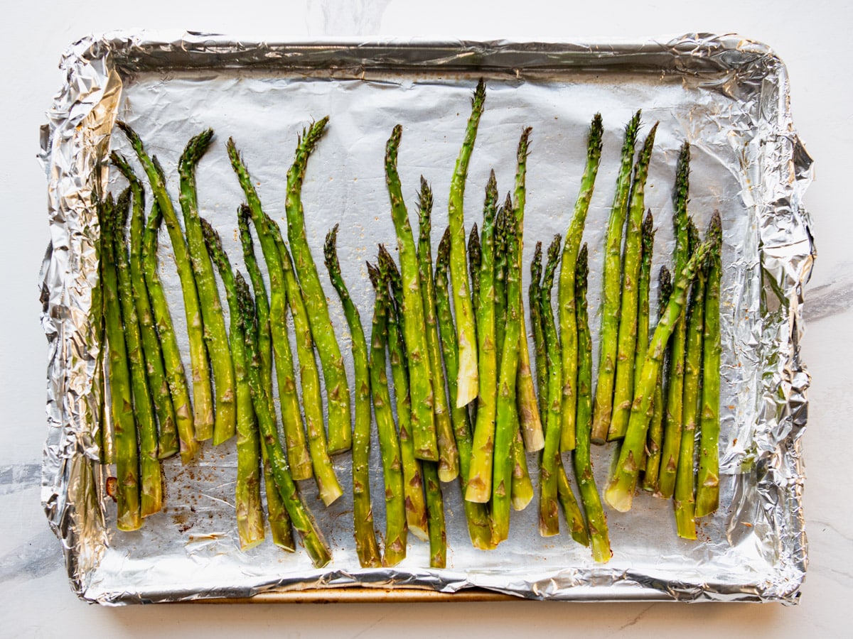 A single sheet pan of oven roasted asparagus spears with lightly blistered tips, grated Parmesan, and lemon wedges on a warm kitchen counter under soft natural light