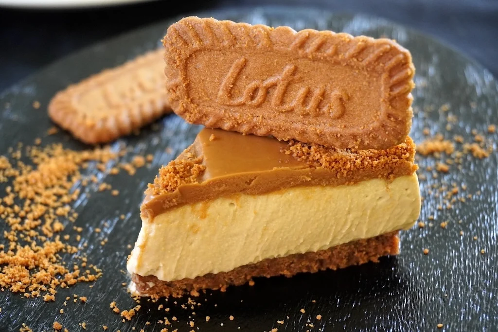 A single slice of Biscoff cheesecake on a small dessert plate, showing a thick cookie crust and a cookie butter swirl on top, set on a warm-toned kitchen table with soft natural light