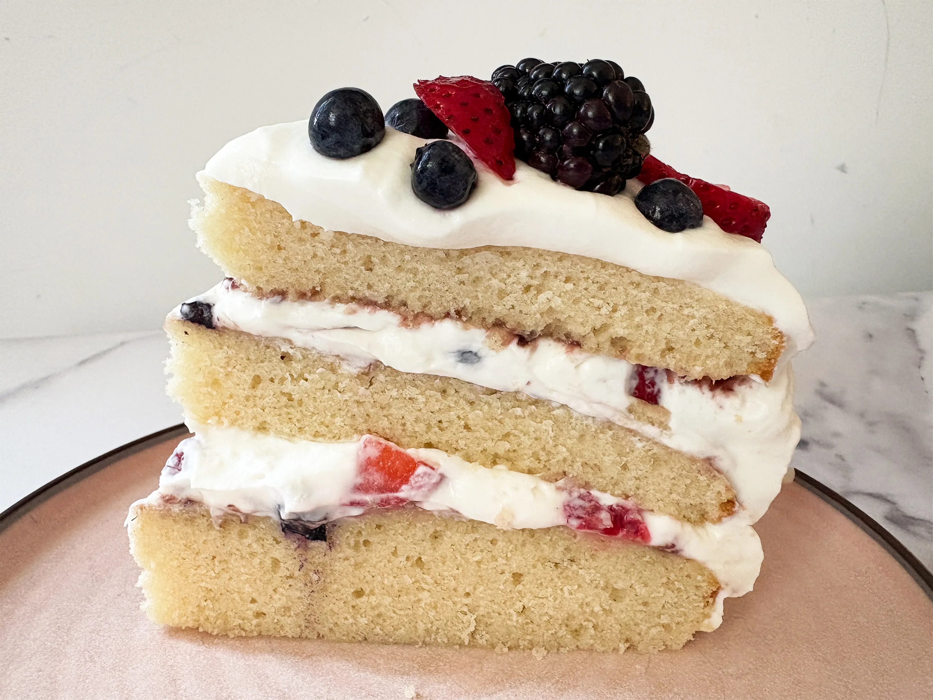 A single slice of Chantilly cake with visible vanilla layers, mascarpone cream, and mixed berries on a dessert plate with a fork beside it