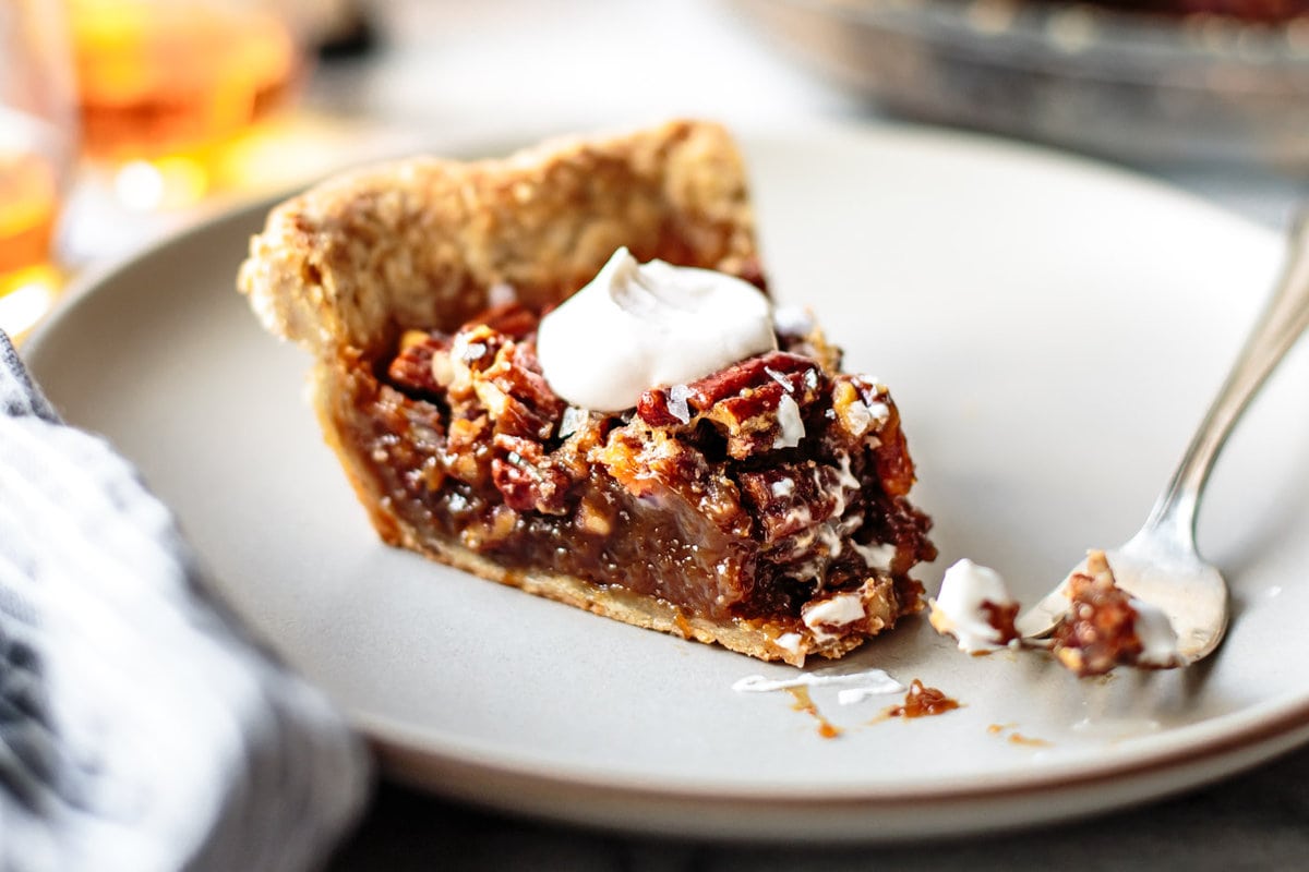 A single slice of bourbon pecan pie on a small plate with a fork, showing glossy toasted pecans and a gooey center, warm kitchen table lighting