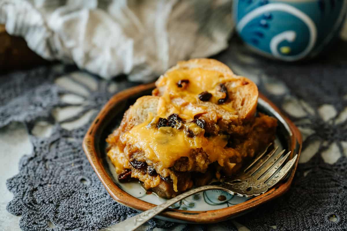 A single slice of capirotada on a small plate with a spoon beside it, showing layers of bread, raisins, nuts, and melted cheese in warm kitchen lighting