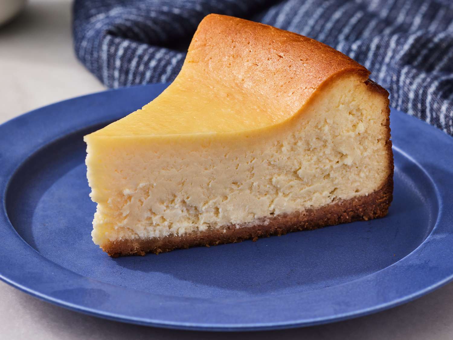 A single slice of classic baked cheesecake with a graham cracker crust on a white dessert plate, creamy interior visible, on a simple wooden table setting with soft natural light