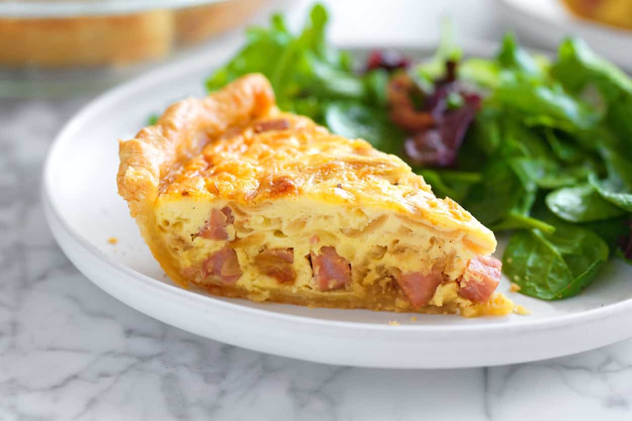 A single slice of classic quiche on a ceramic plate with a golden flaky crust and creamy egg filling, set on a sunlit kitchen table
