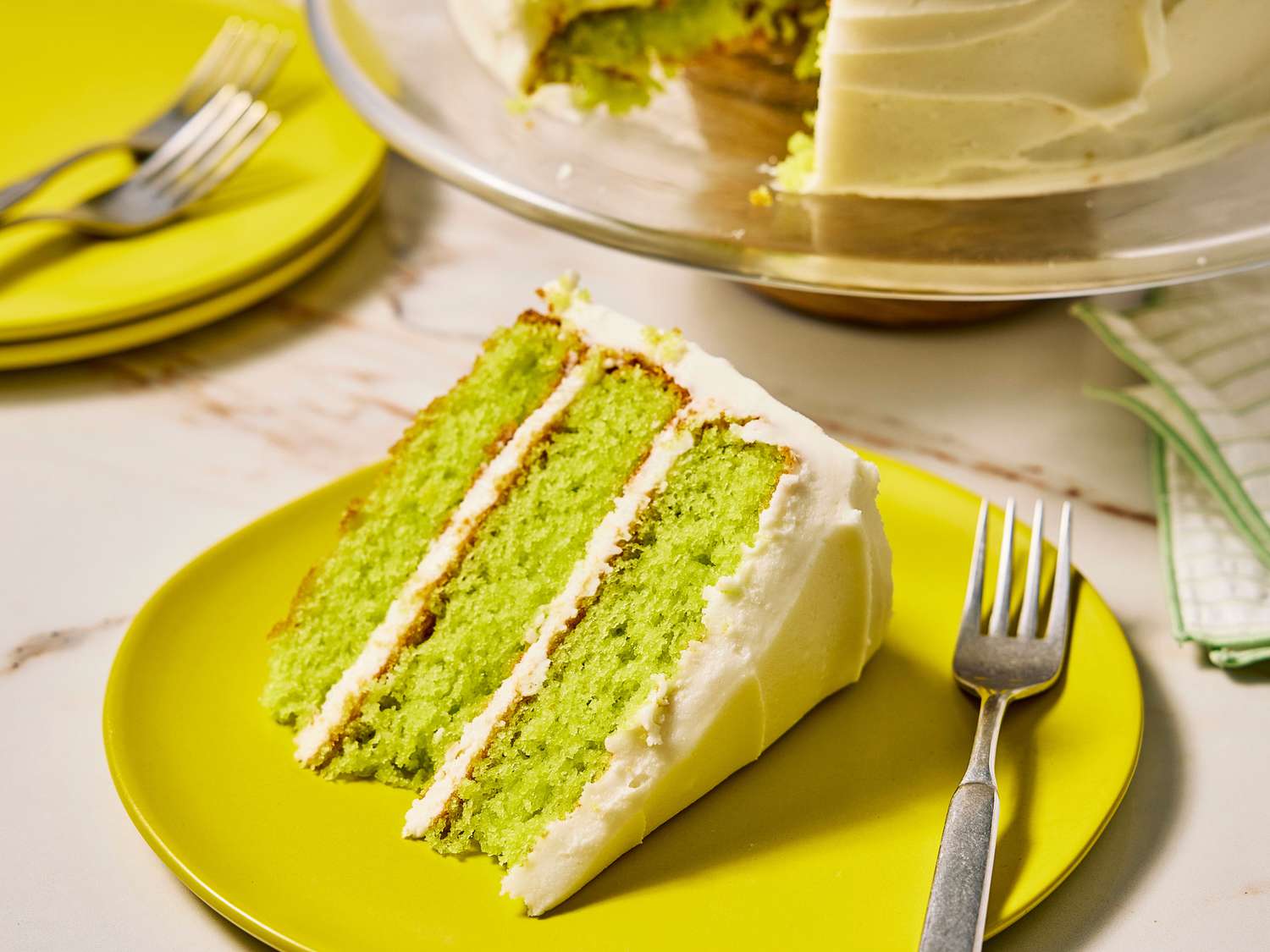 A single slice of key lime cake with cream cheese frosting on a small plate with a fork beside it on a linen napkin