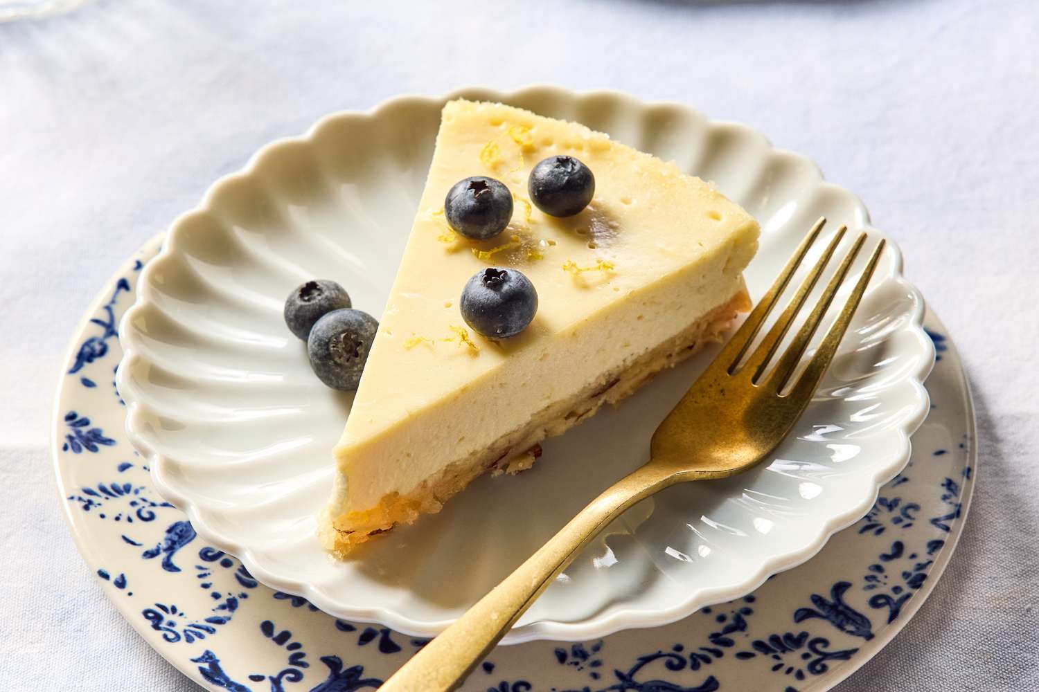 A single slice of ricotta cheesecake on a small ceramic plate with a pale golden top, a crisp cookie crust, and a few lemon zest curls on the side, set on a light stone kitchen counter in soft natural window light