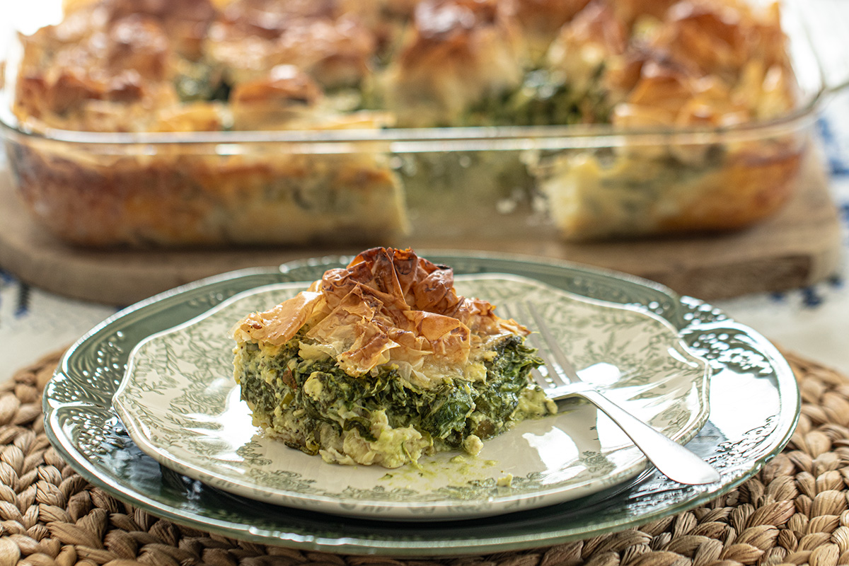 A single slice of spinach and feta filo pie on a ceramic plate with visible flaky layers and a small pile of herbs on the side