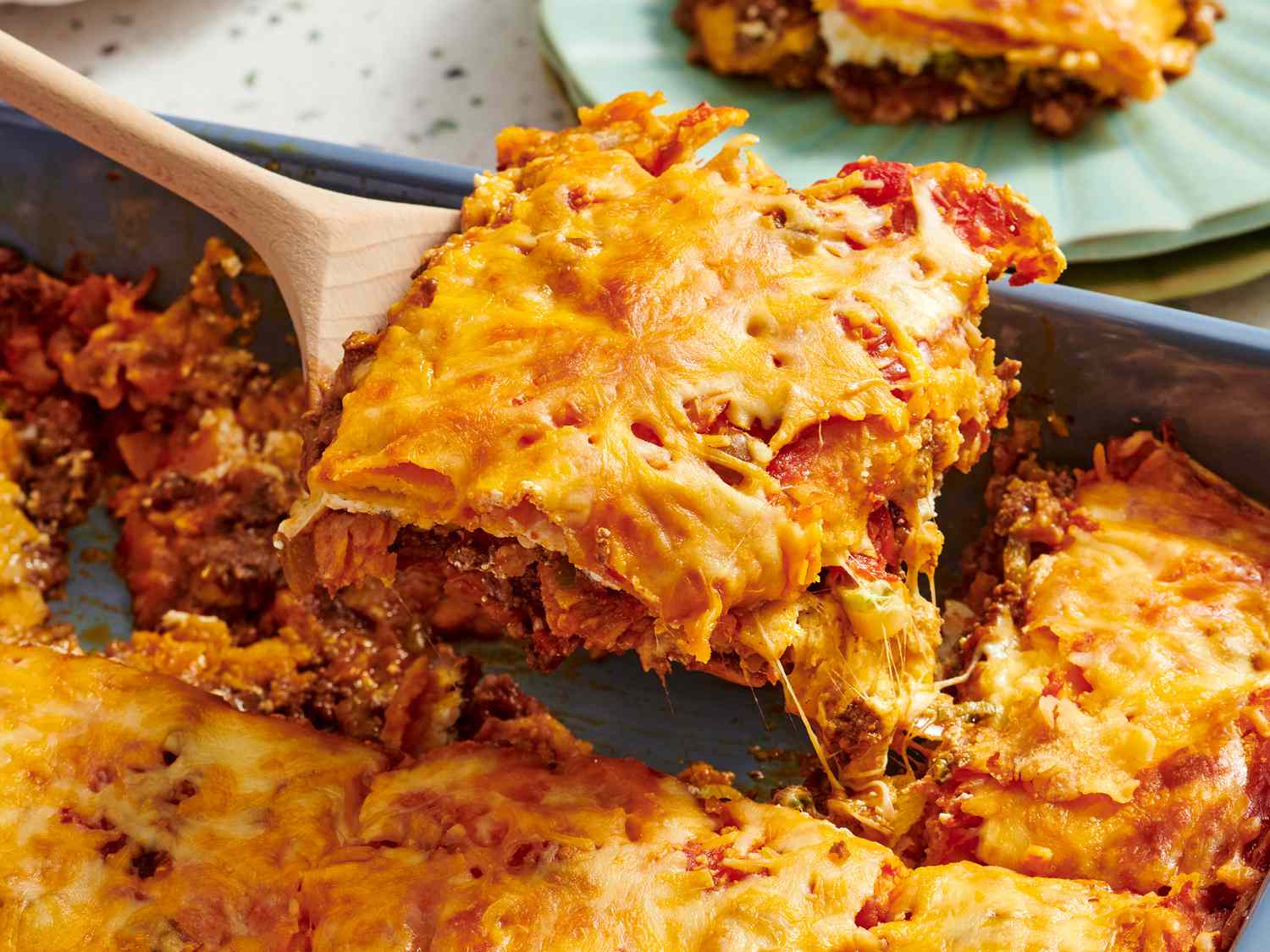 A single square slice of Mexican lasagna on a plate with melted cheese and visible tortilla layers, with a fork beside it on a simple dining table