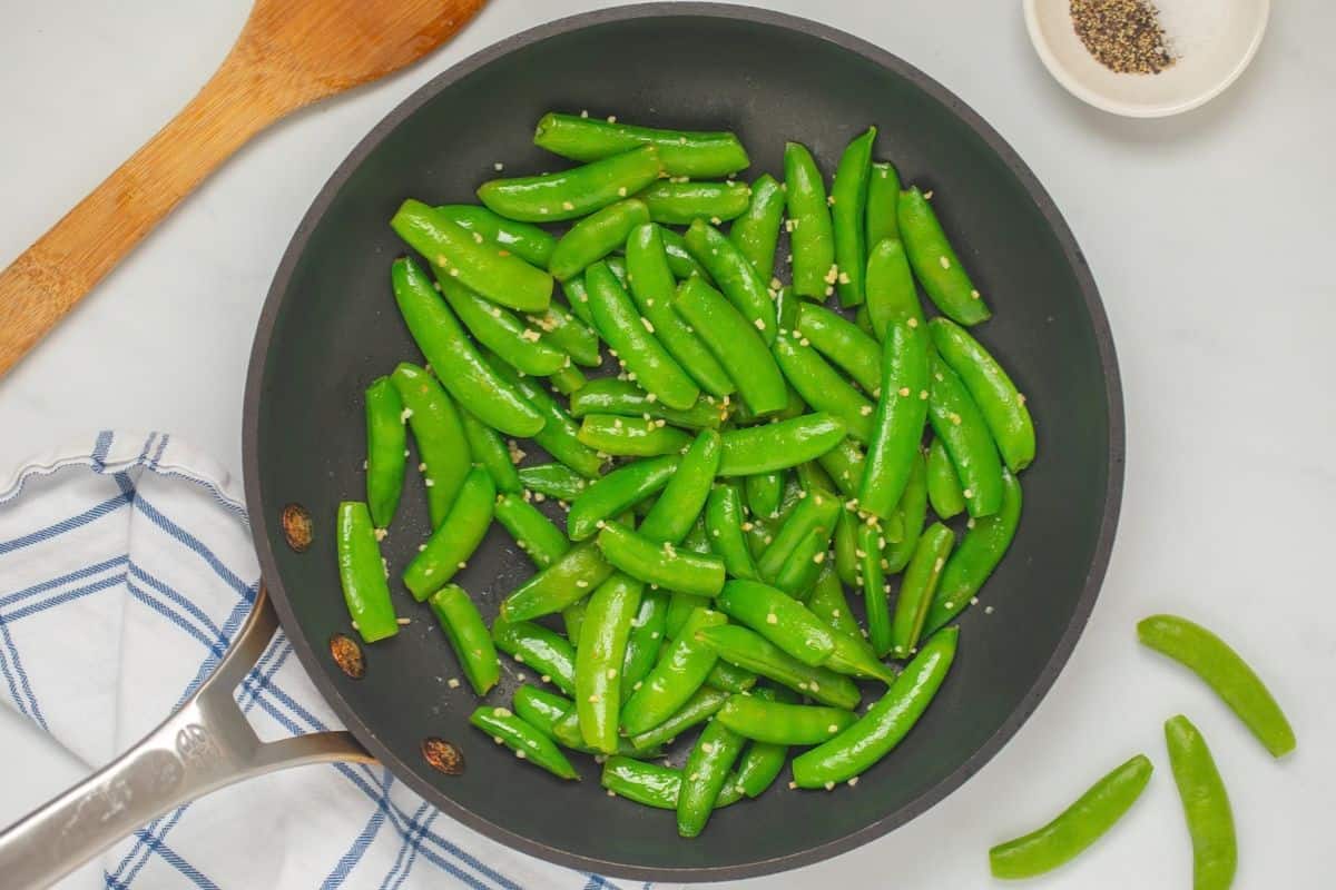 A skillet filled with bright green snap peas tossed in glossy garlic butter with visible bits of minced garlic and lemon zest, set on a warm kitchen countertop