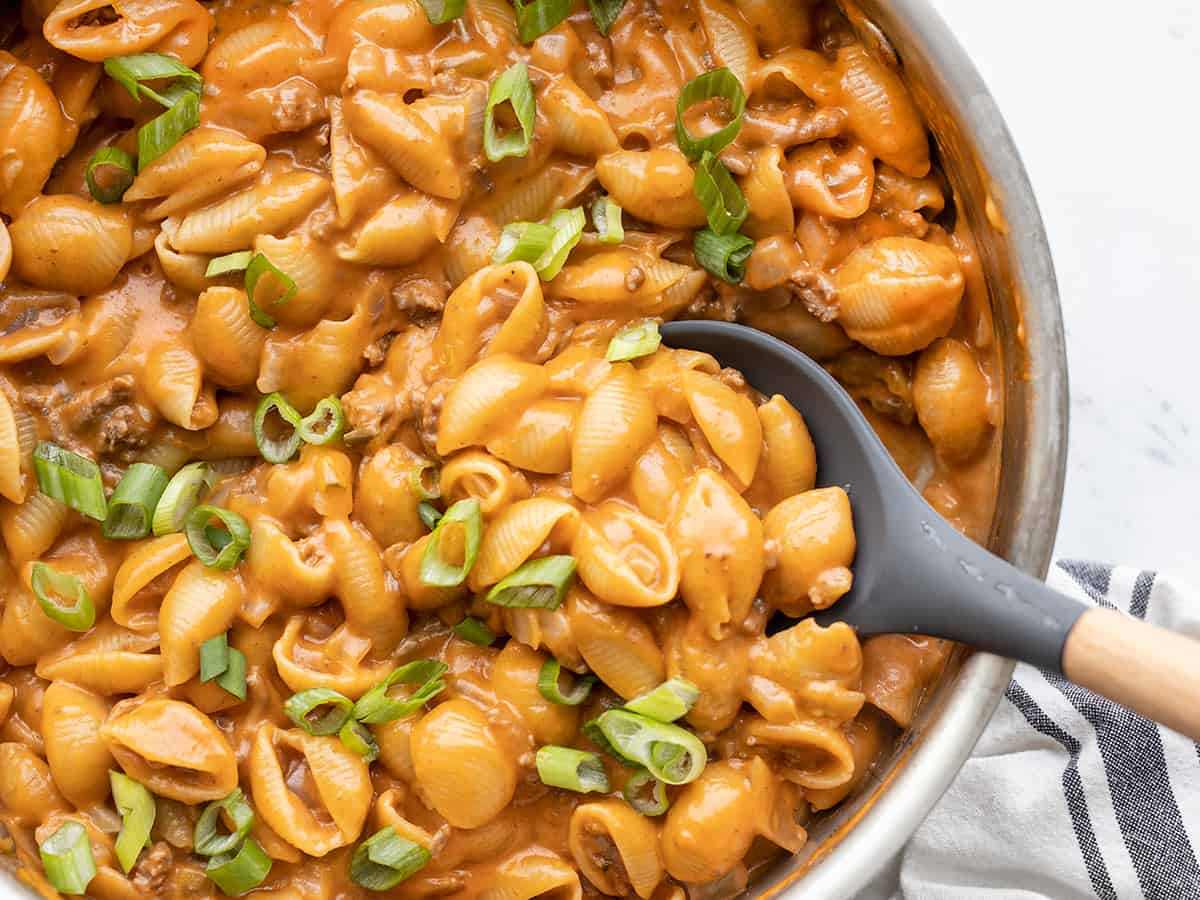 A skillet of creamy cheeseburger macaroni topped with melted cheddar and sliced green onions on a warm kitchen counter