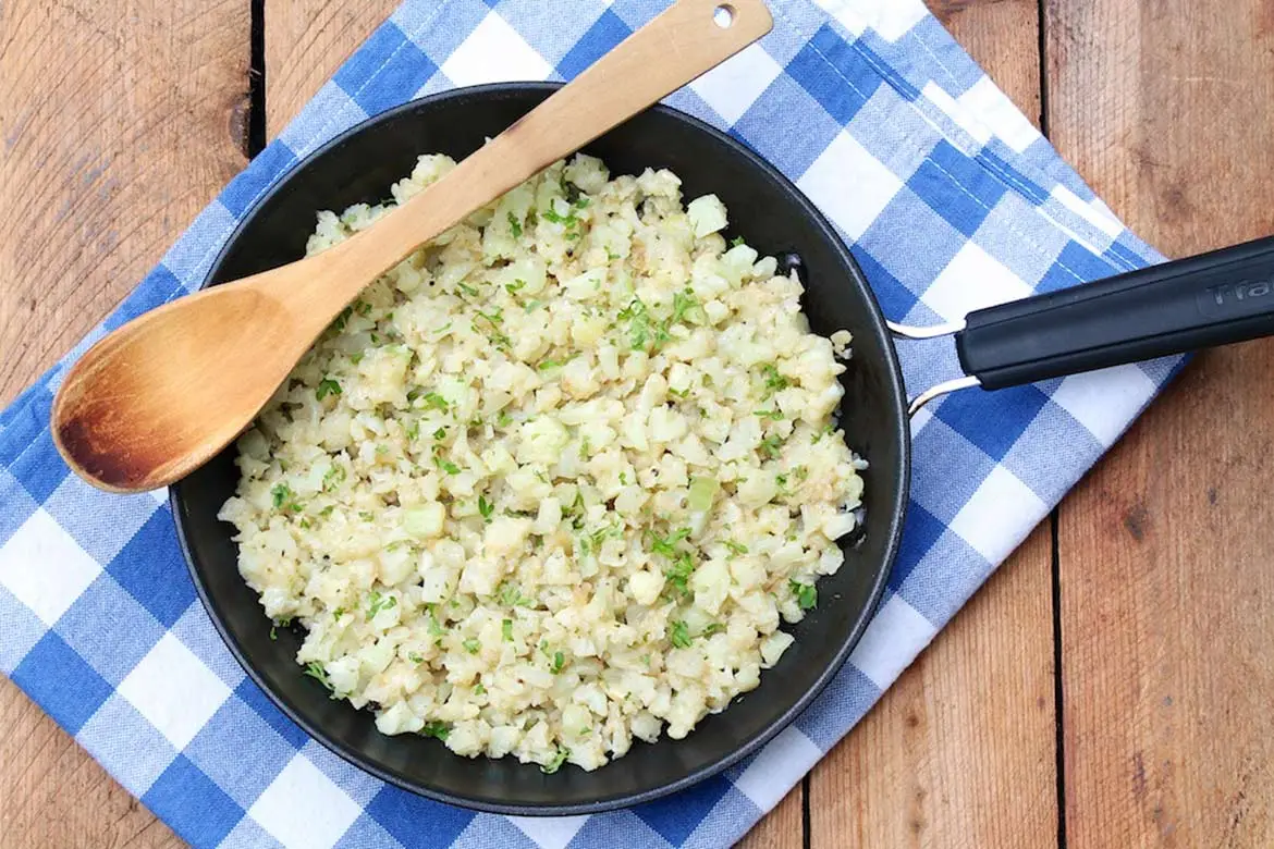 A skillet of crispy-edged cauliflower rice tossed with garlic and Parmesan, finished with chopped parsley and lemon wedges on a warm kitchen counter