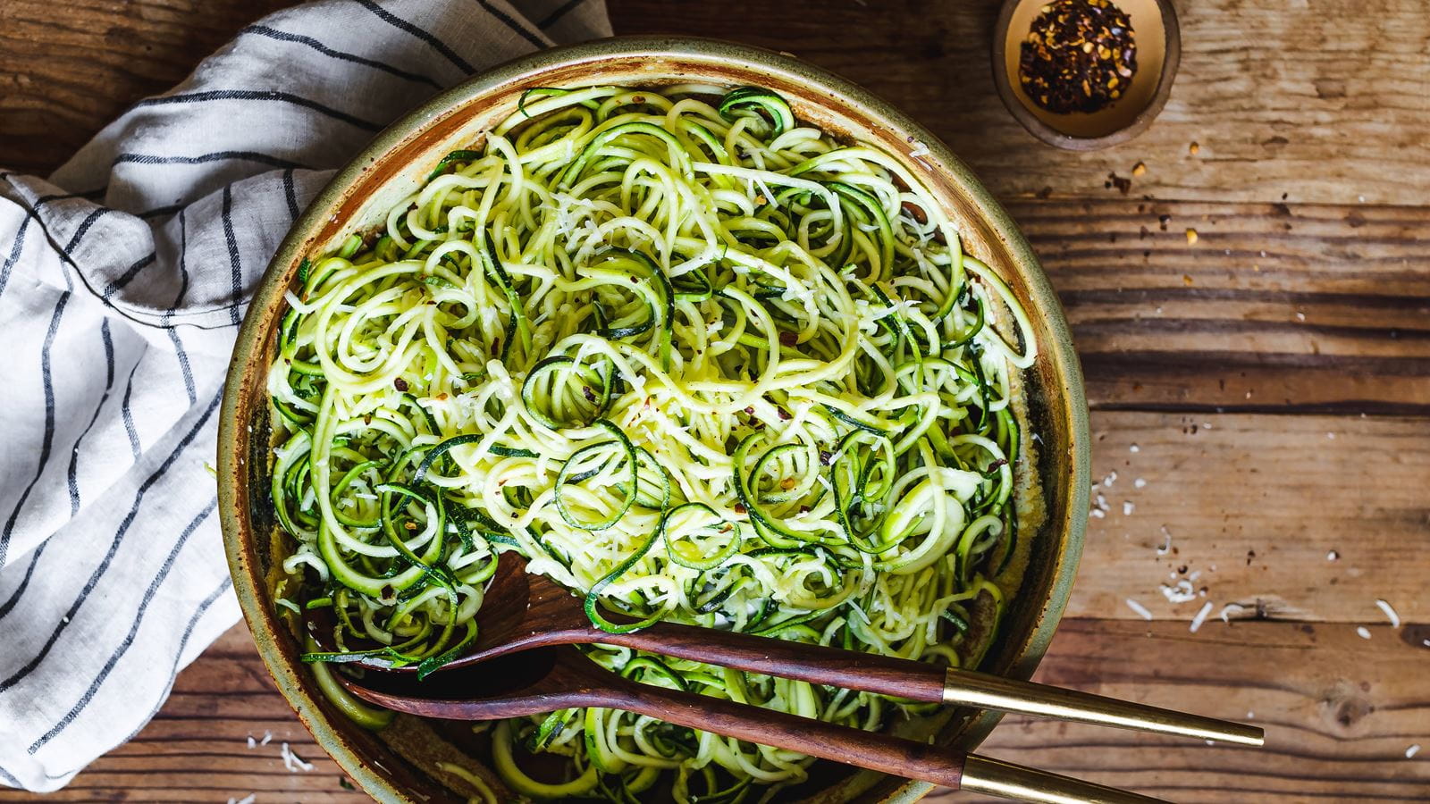 A skillet of garlic Parmesan zucchini noodles tossed with lemon zest and black pepper, with a wooden spoon resting on the pan in a warm home kitchen