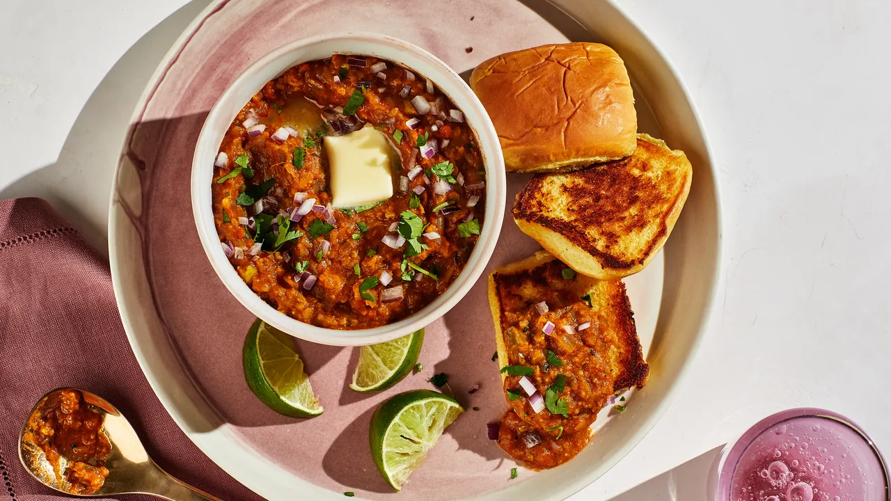 A skillet of glossy, orange-red pav bhaji topped with a pat of butter, chopped cilantro, and a lemon wedge on the side, with toasted rolls nearby on a warm kitchen counter
