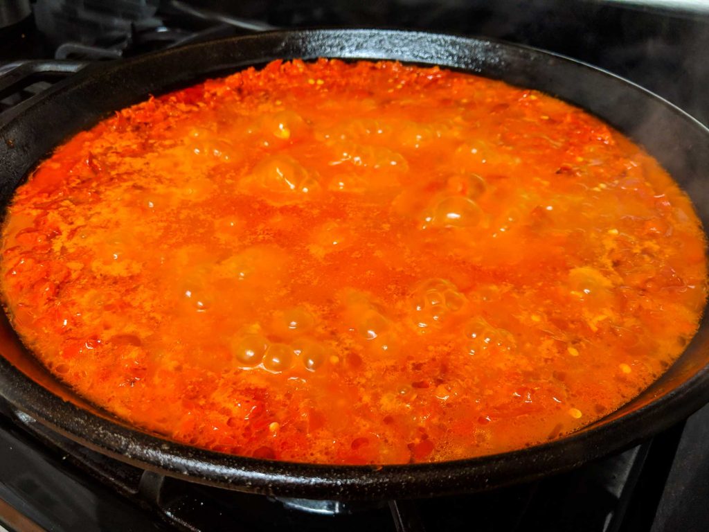 A skillet on a stove with red sambal being stirred with a wooden spoon, showing glossy chili paste bubbling gently