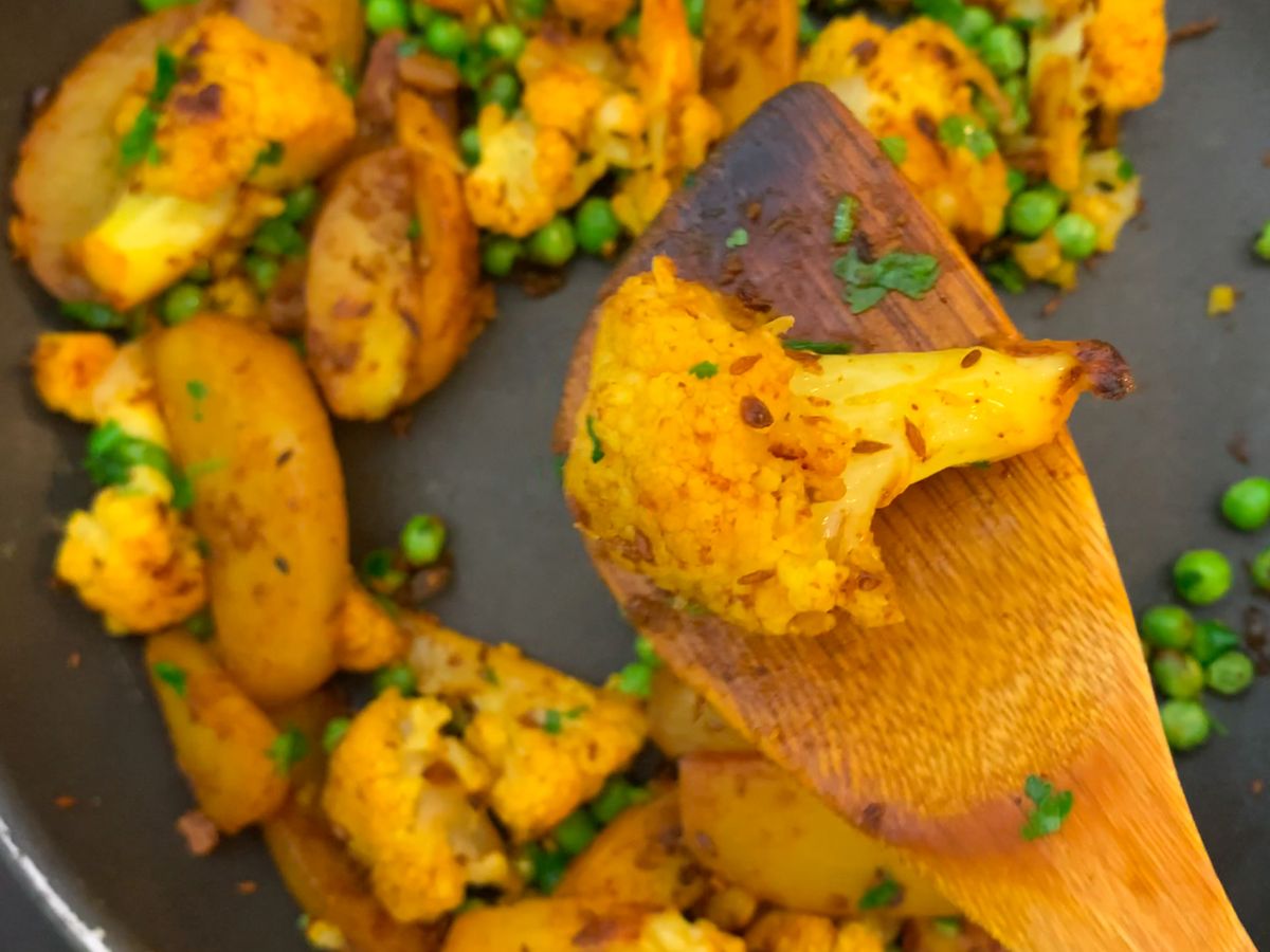 A skillet on a stovetop with aloo gobi simmering, potatoes and cauliflower coated in yellow spices while steam rises