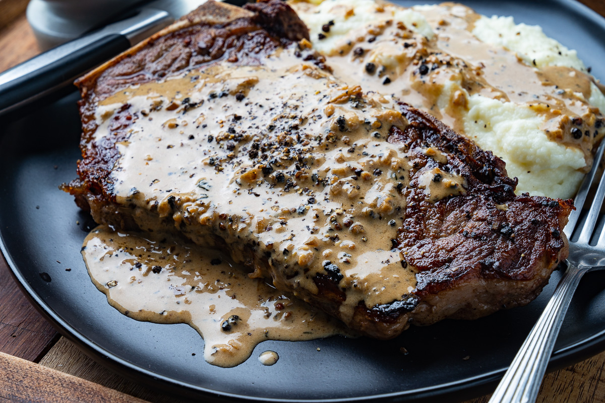 A skillet on a stovetop with peppercorn-crusted steaks searing while butter foams around them