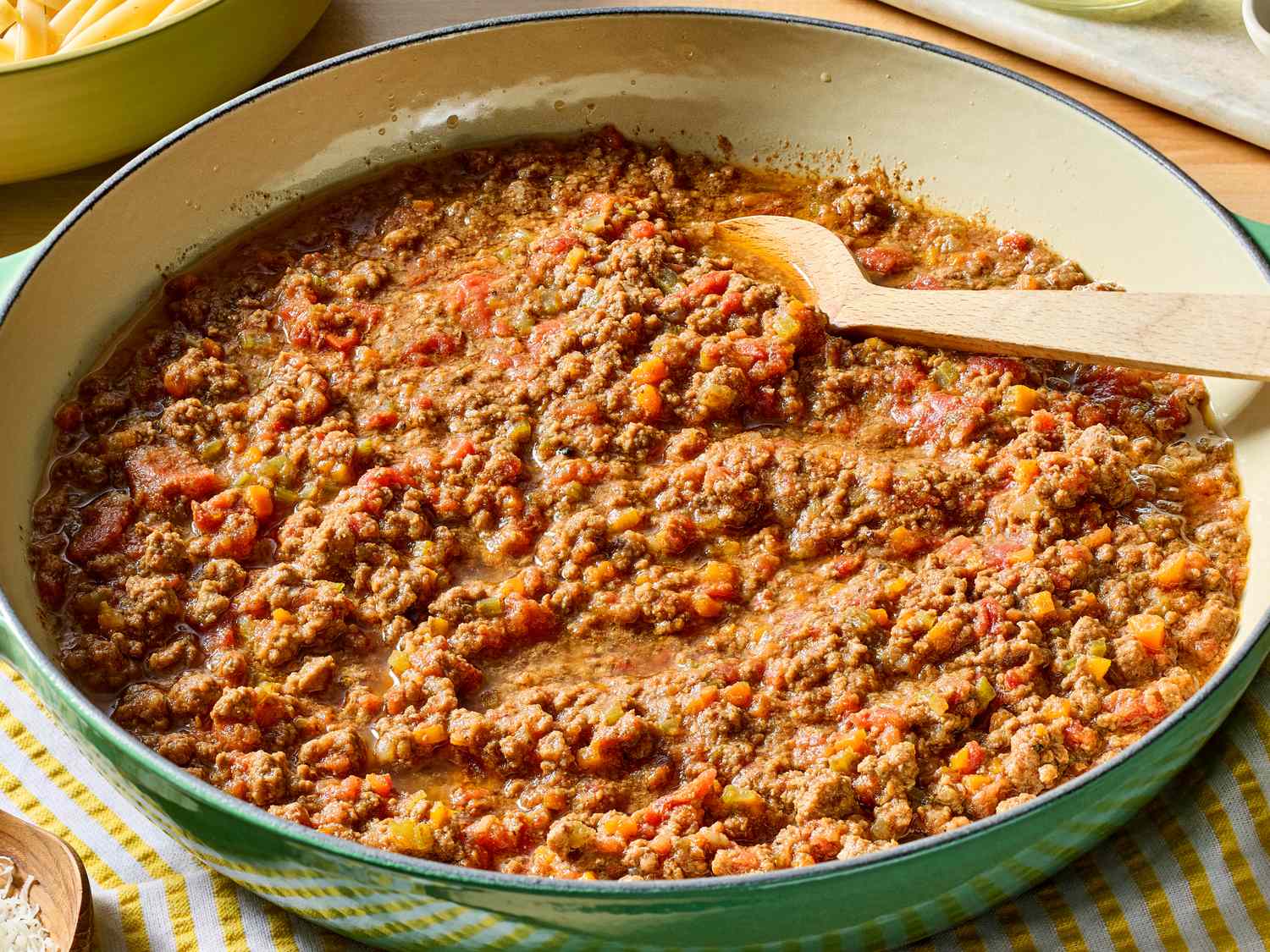A skillet on the stove with browned ground beef and marinara sauce simmering, with a wooden spoon resting on the edge