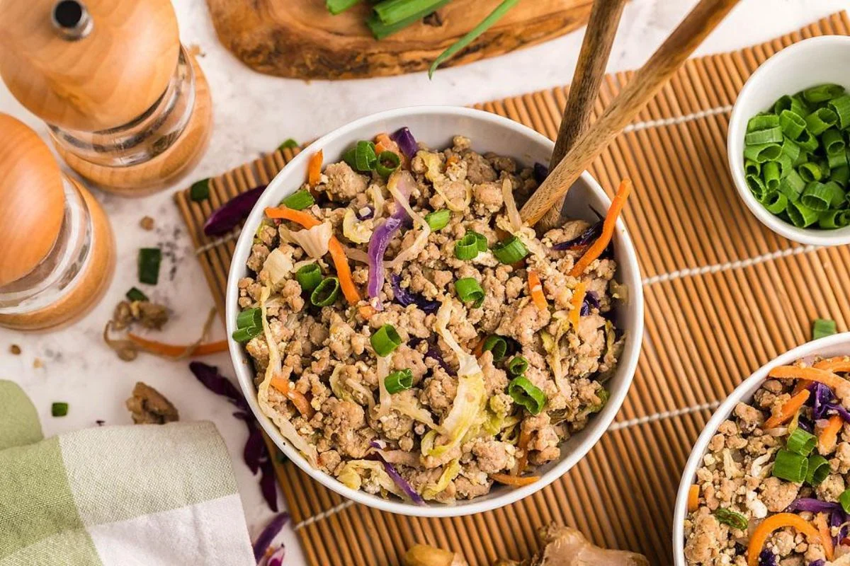 A skillet on the stove with browned ground pork, shredded cabbage, and carrots being stirred together into an egg roll filling
