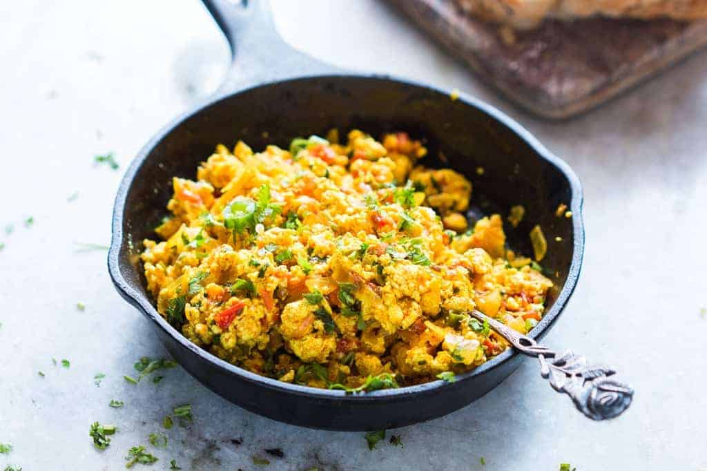 A skillet on the stove with paneer bhurji being stirred by a wooden spoon, showing steam rising and glossy onion-tomato masala coating the crumbles