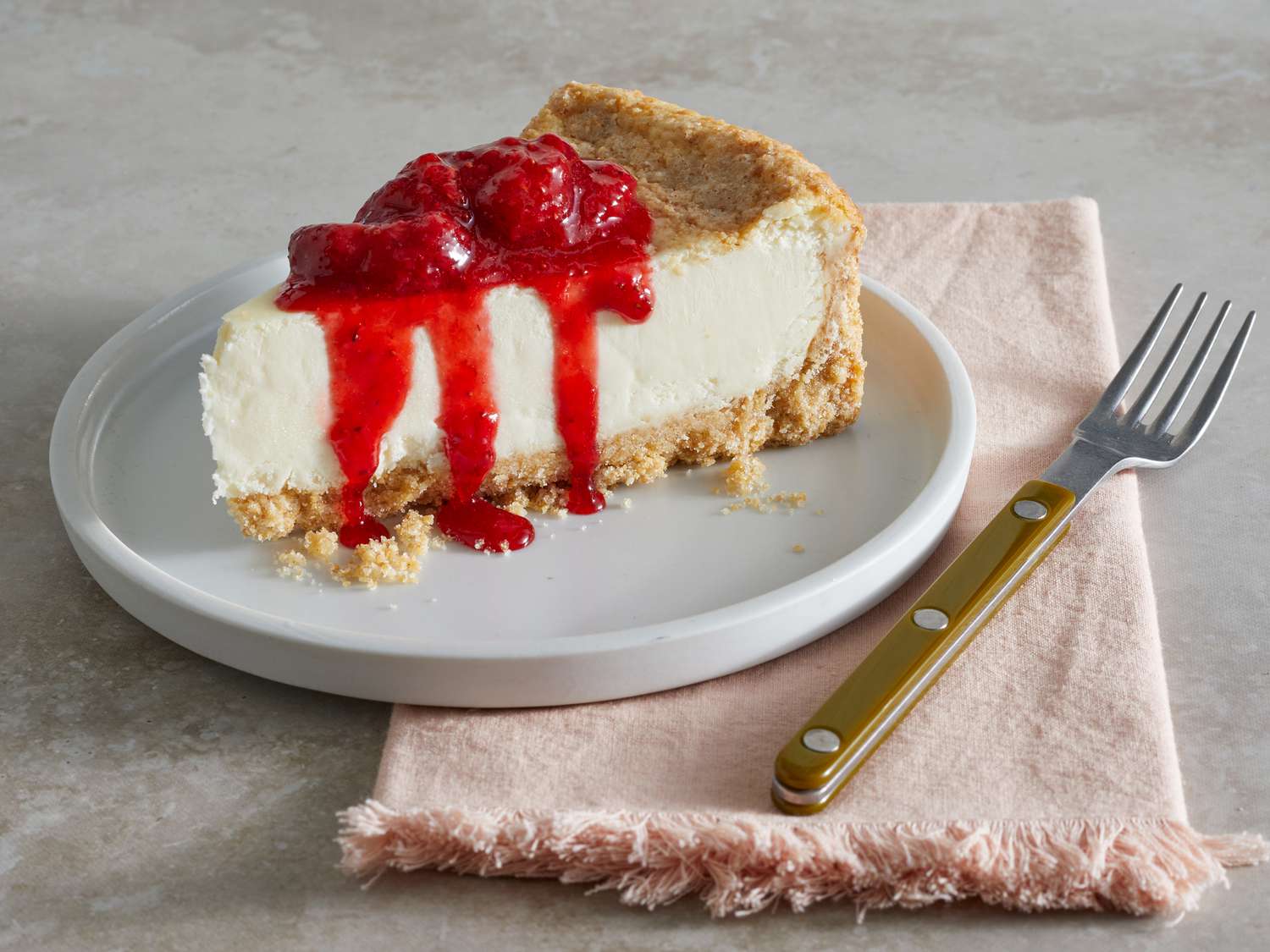 A slice of classic cheesecake topped with a thick, glossy strawberry glaze dripping slightly down the sides on a white plate on a sunlit kitchen table