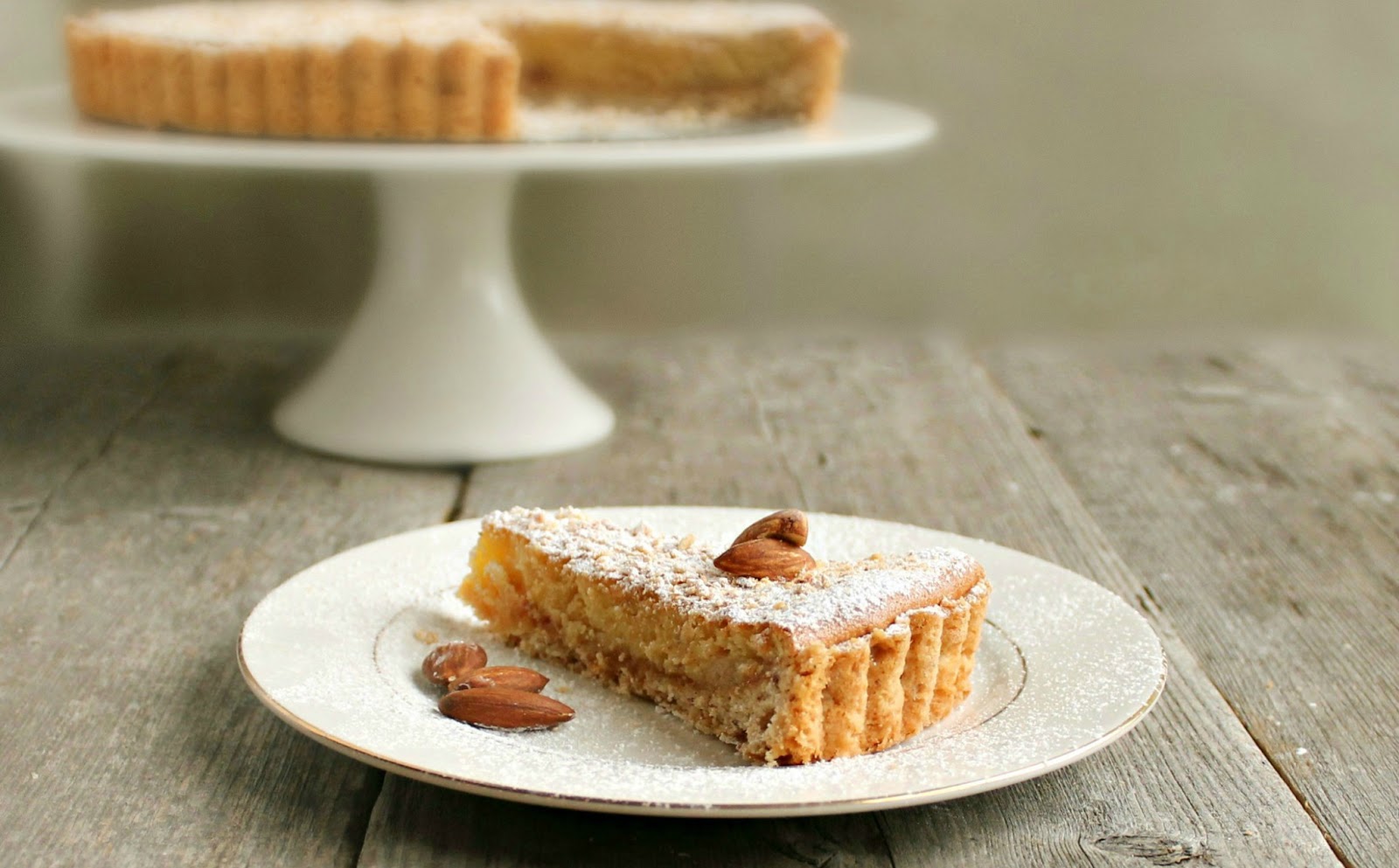 A slice of fruit tart with golden baked frangipane filling on a ceramic plate, crumbs scattered on a warm kitchen table