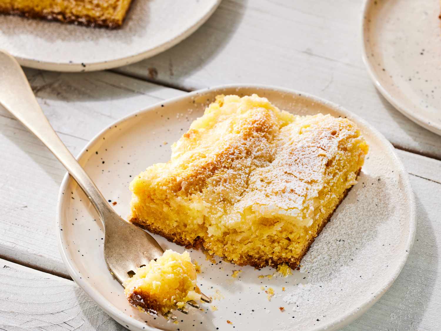 A slice of ooey gooey butter cake being dusted with powdered sugar over a parchment-lined tray