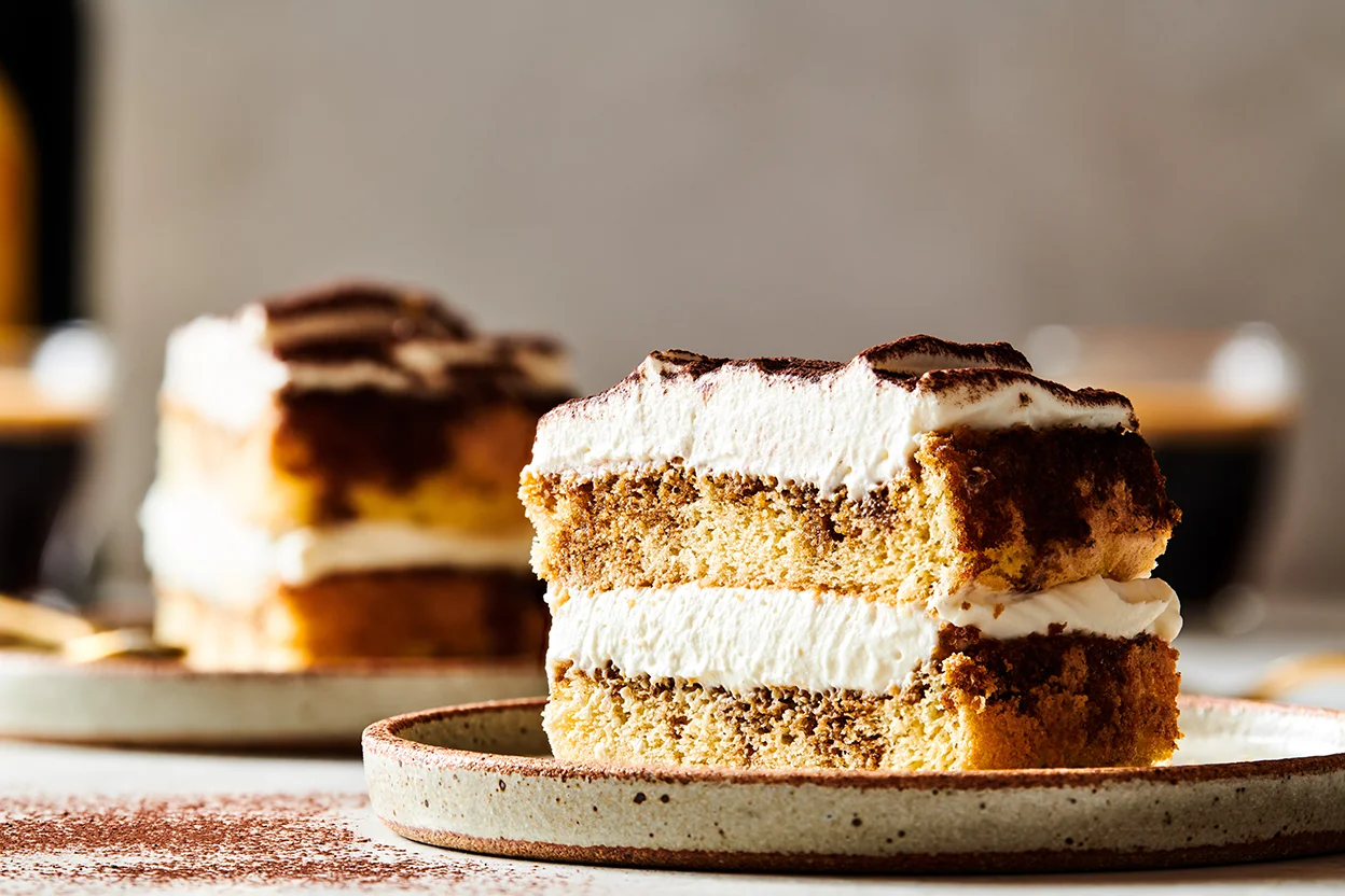 A slice of tiramisu layer cake on a small dessert plate, showing vanilla sponge layers soaked with coffee and thick mascarpone cream, finished with a dusting of cocoa on top, on a warm-toned kitchen table