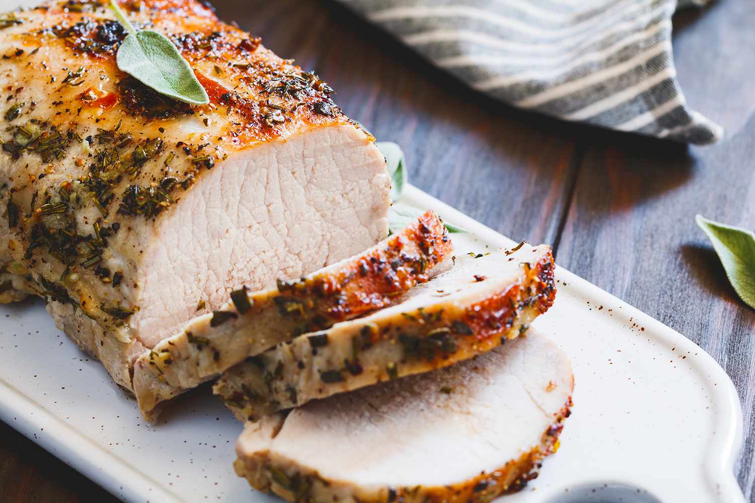 A sliced boneless pork loin roast on a white platter with a golden brown crust, rosemary sprigs, and pan juices, set on a warm wood kitchen table