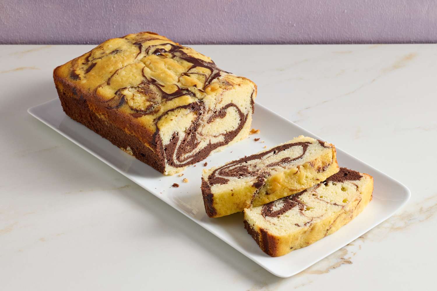 A sliced marble loaf cake showing dramatic vanilla and chocolate swirls, resting on a wooden cutting board with a small knife and a few crumbs in warm kitchen light