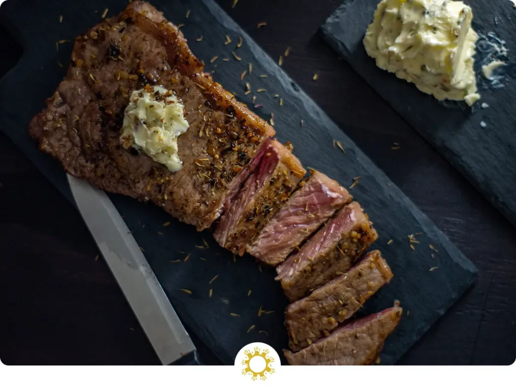 A sliced medium-rare ribeye steak on a white plate with melted garlic herb butter and flaky salt, served with a small pile of arugula on a wooden table