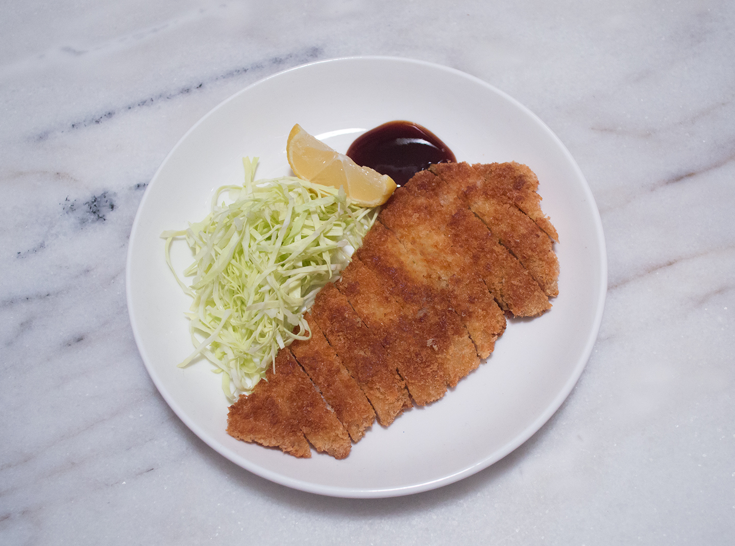 A sliced pork katsu cutlet with a crisp golden panko crust on a plate with shredded cabbage, a lemon wedge, and a small ramekin of katsu sauce on a wooden table