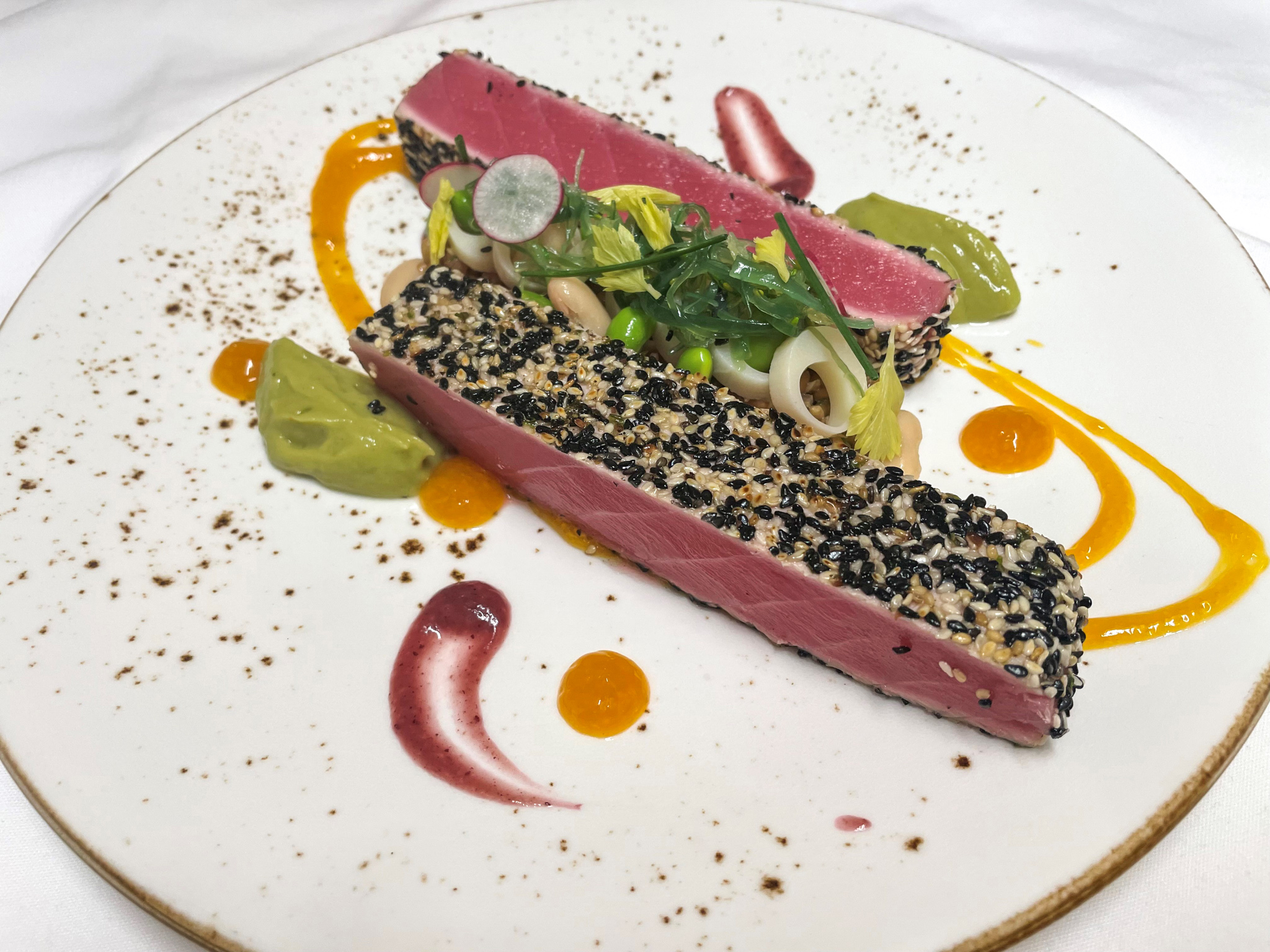A sliced sesame-crusted seared ahi tuna steak fanned on a white plate with a small bowl of soy-lime dipping sauce on a wooden table
