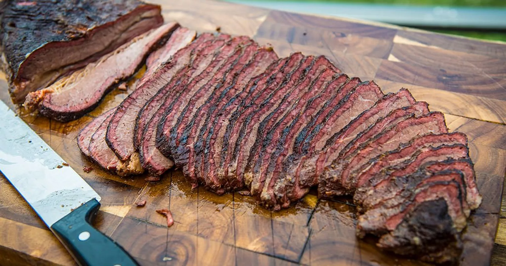 A sliced smoked beef brisket on a wooden cutting board with a dark peppery bark and visible smoke ring, juices glistening under warm kitchen lighting