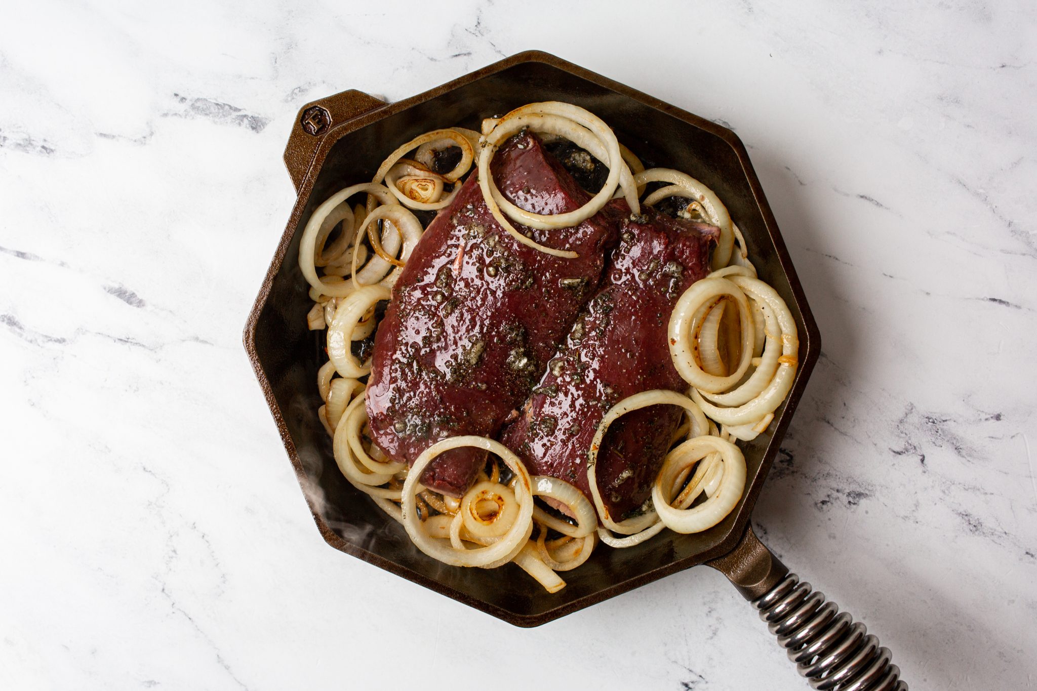 A sliced venison steak cooked to medium-rare on a white plate with melted garlic herb butter pooling on top, a rosemary sprig, and a cast-iron skillet nearby on a warm kitchen counter