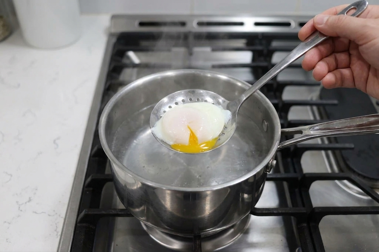 A slotted spoon lifting a freshly poached egg from simmering water in a stainless steel saucepan on a home stove