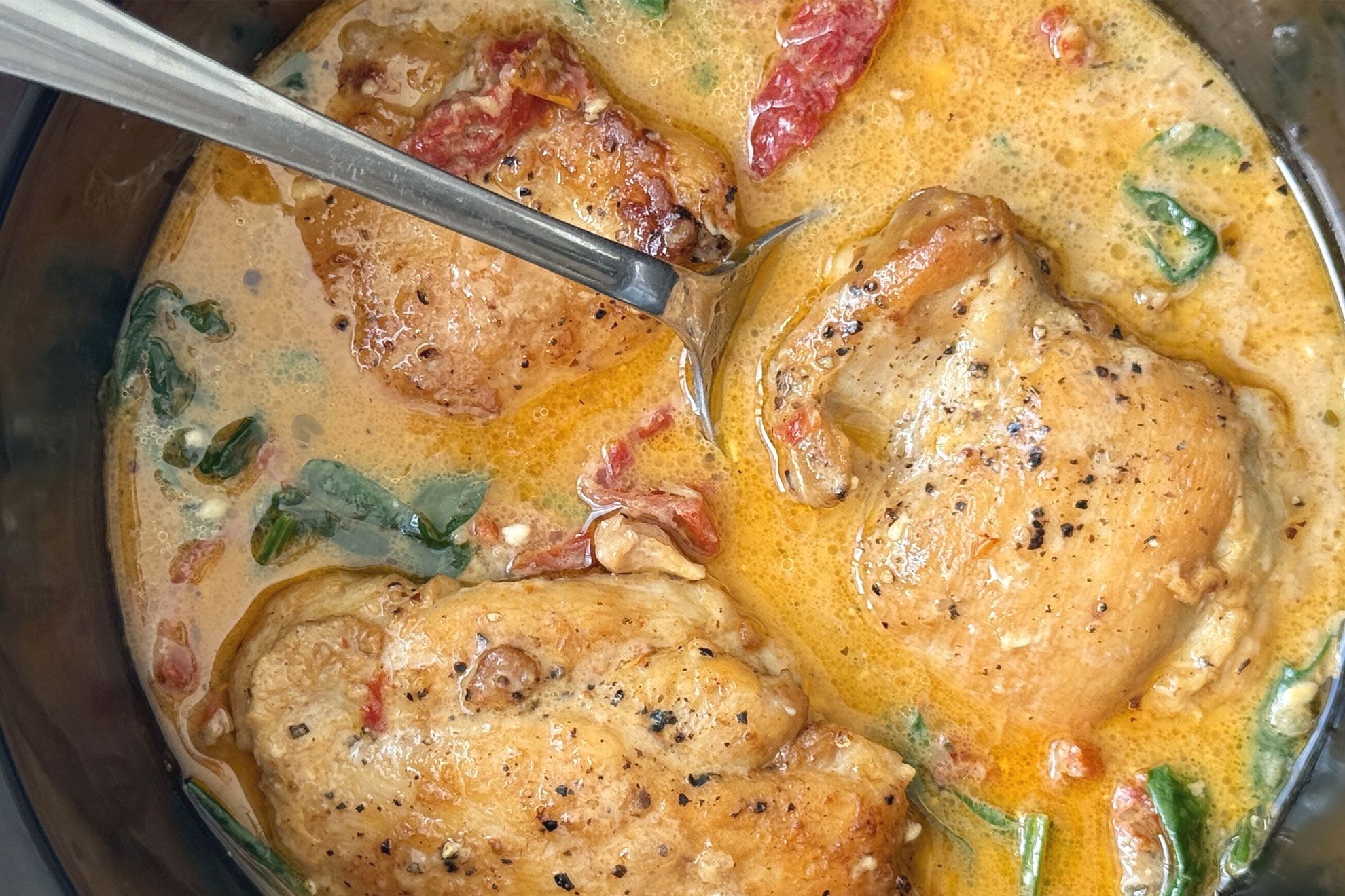 A slow cooker filled with creamy Marry Me chicken sauce with sun-dried tomatoes and herbs, with a serving spoon lifting tender chicken, warm kitchen lighting